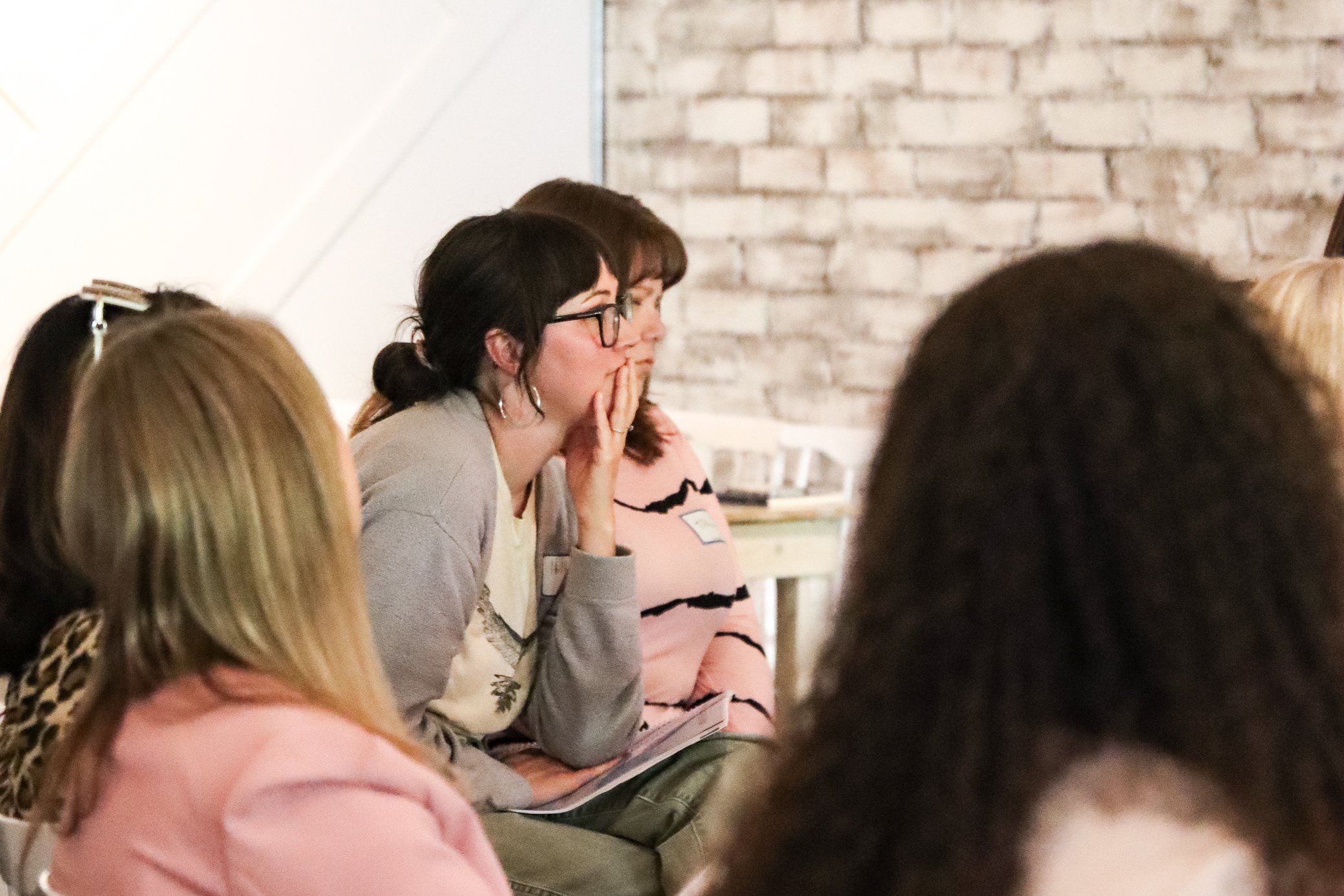 Group of women sitting closely together, listening attentively during a meeting or presentation, with some taking notes, against a brick wall background.