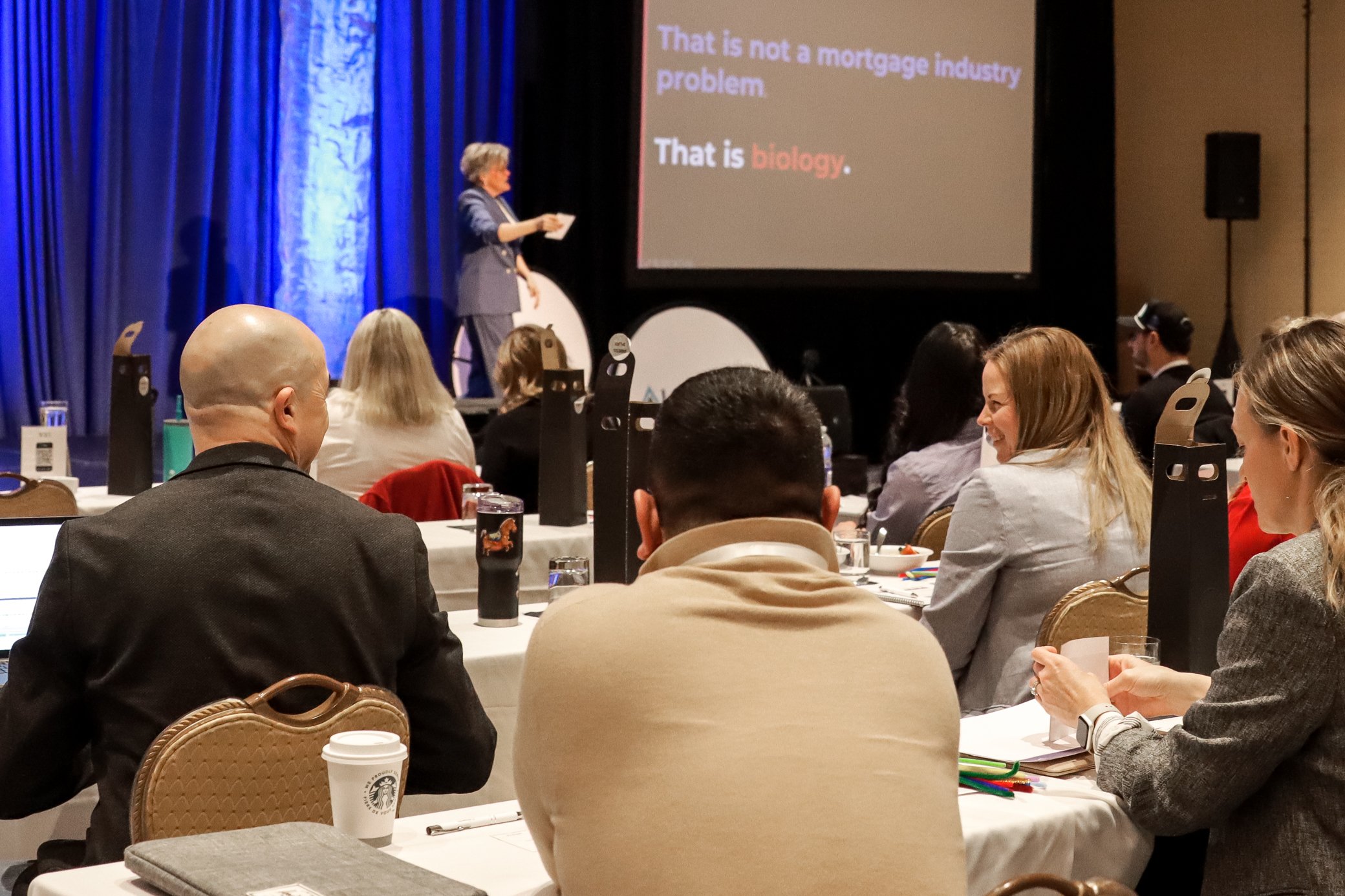 A woman in a blue suit giving a presentation at a conference, standing near a large screen with a slide that reads, 'That is not a mortgage industry problem. That is biology.' The audience is seated at round tables, paying attention to the speaker.