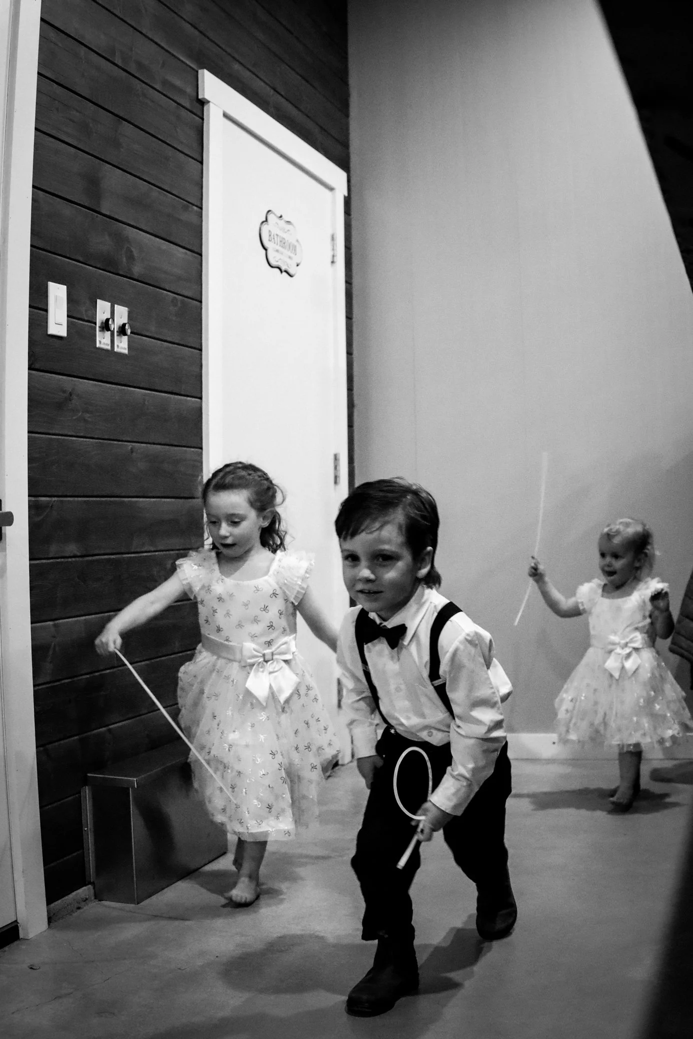 Children dressed in formal attire, including girls in dresses and boys in suits, holding ribbons and participating in a dance or parade indoors, near a door labeled "Bathroom" with wood-paneled walls.