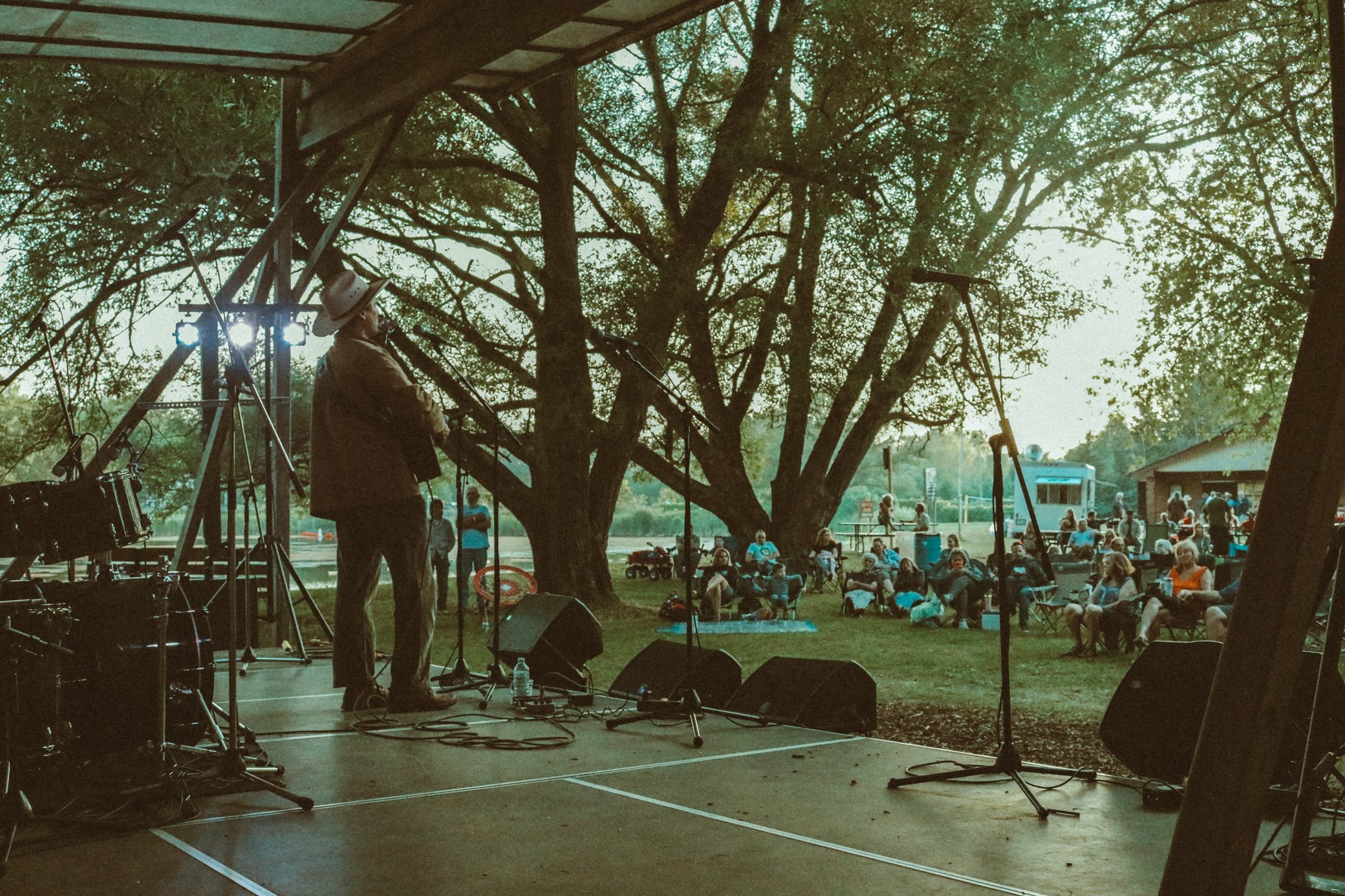 A musician on stage performing at an outdoor concert with a large audience seated in front of the stage during daytime, surrounded by trees.