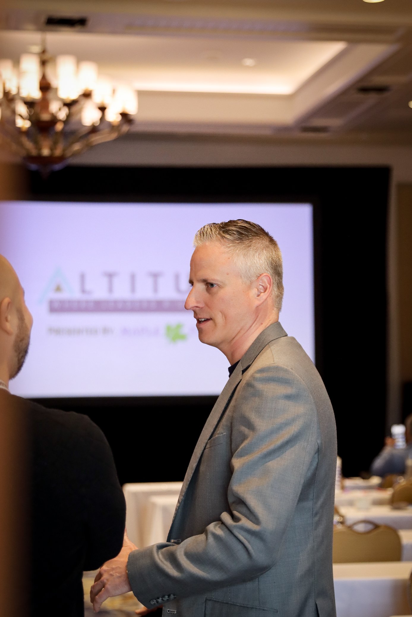 A man with blonde hair and wearing a gray suit is engaged in conversation with another person in a black shirt, indoors at a formal event or conference. In the background, there is a large screen displaying a partly visible logo and text, along with 