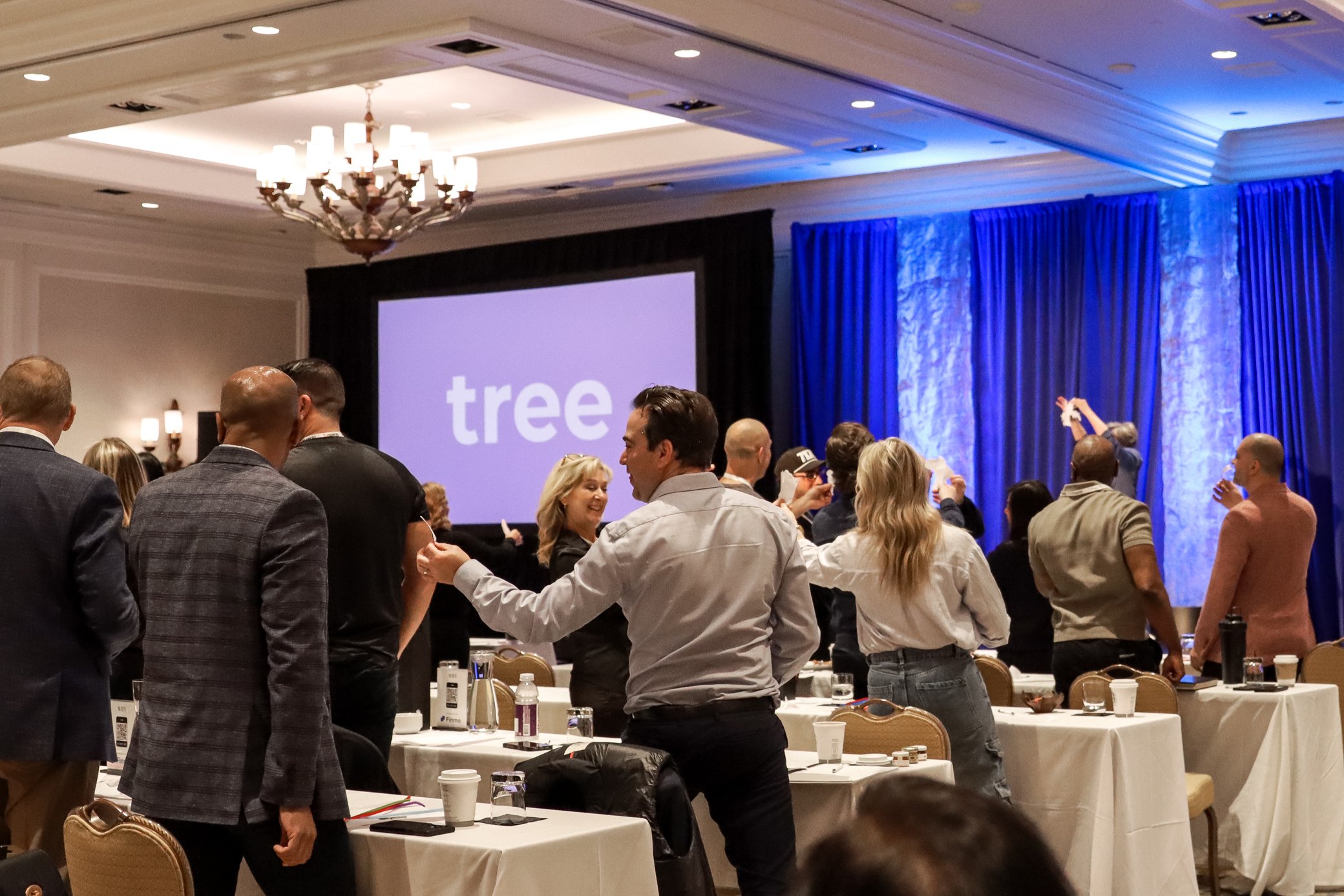 A large conference room filled with people standing and talking, with tables and chairs arranged around the room. A large projection screen displays the word "tree," and blue curtains hang along the side. The room has a high ceiling with chandeliers 