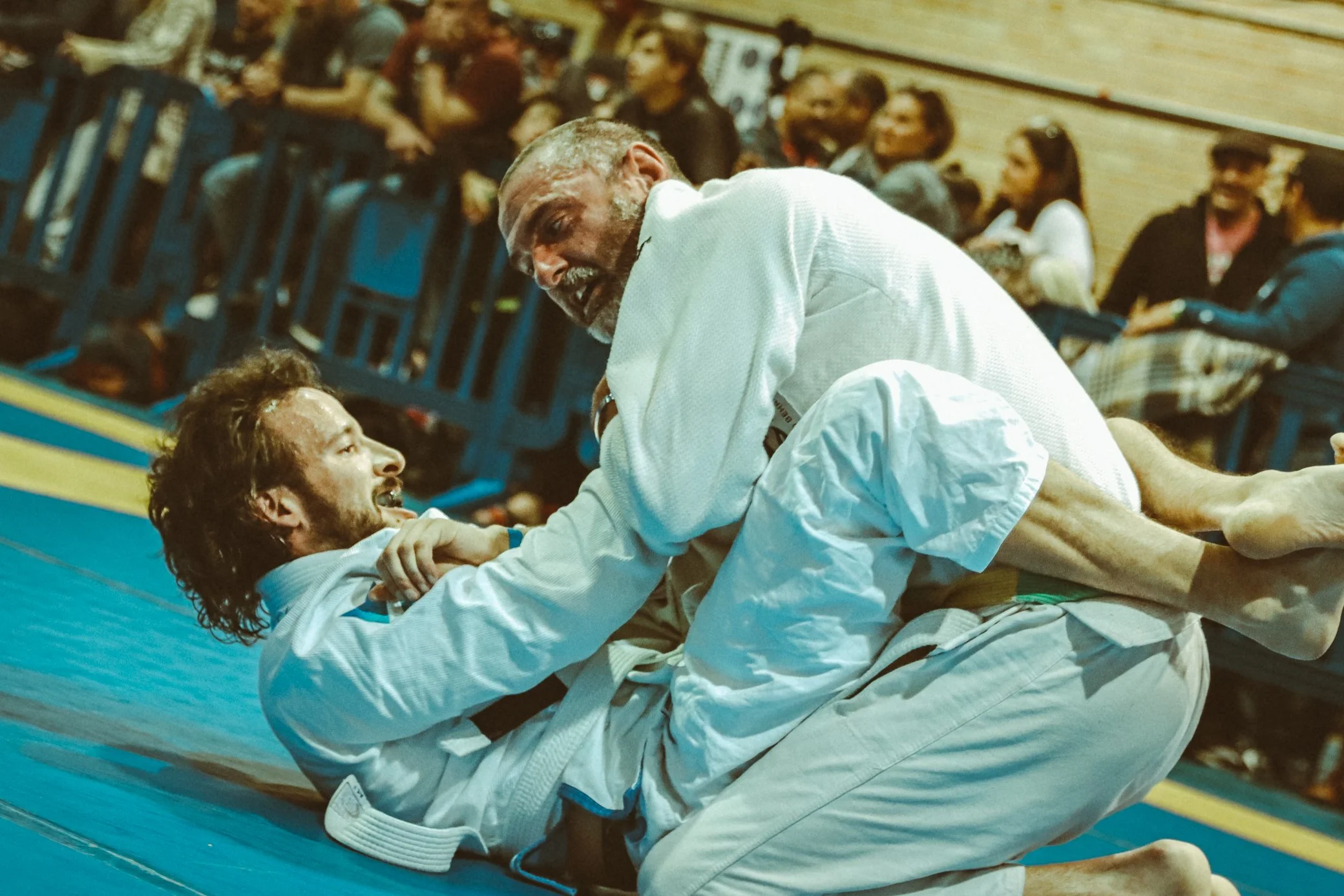 Two men wearing white martial arts uniforms practicing Brazilian Jiu-Jitsu on a blue mat in a gymnasium. One man is on his back while the other is leaning over him, engaging in a grappling move. Several spectators are seated and watching in the backg