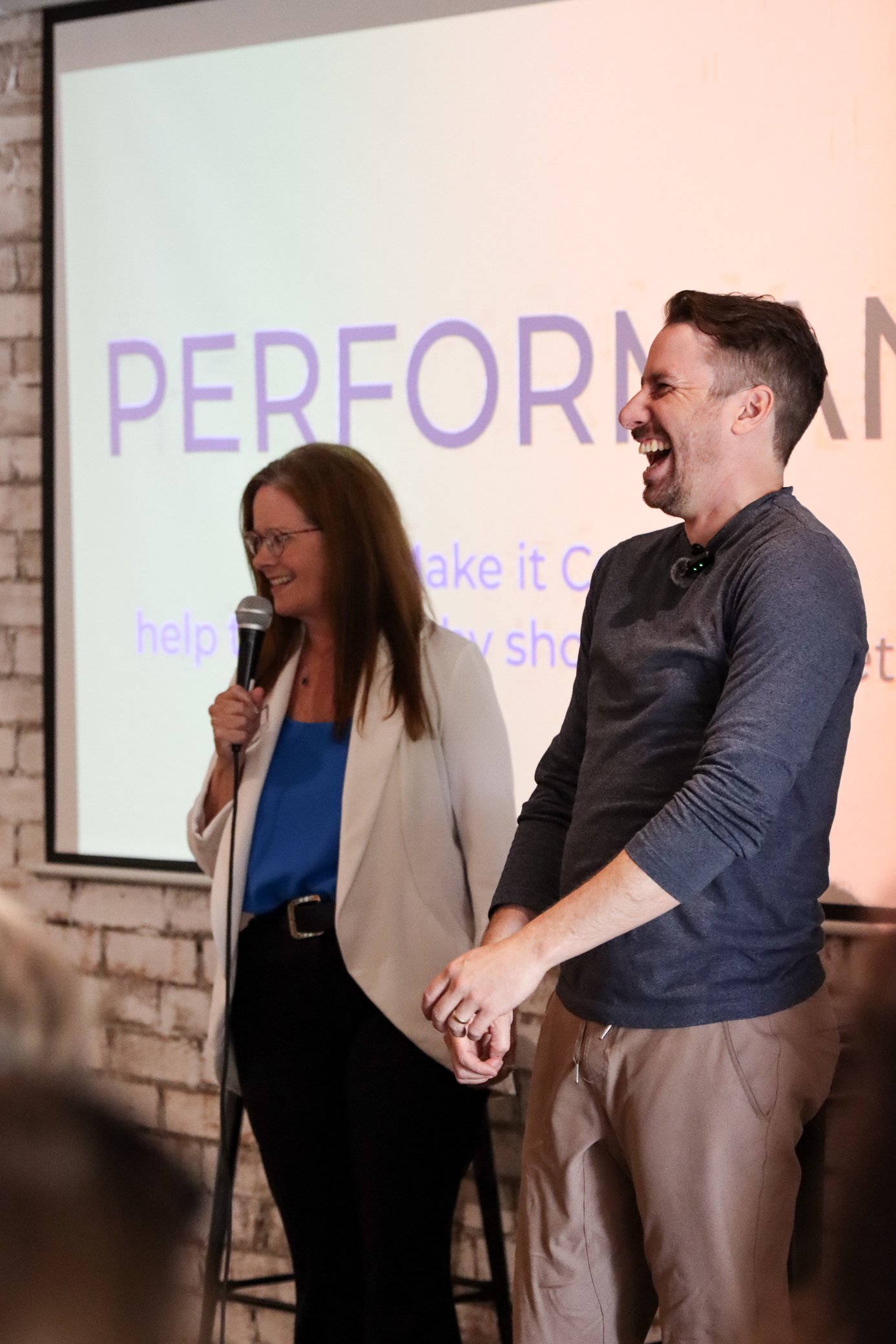 Two people are standing on a stage or in front of a presentation screen, laughing. The woman on the left is holding a microphone, wearing glasses, a white blazer, and a blue top. The man on the right is wearing a dark long-sleeve shirt and light-colo