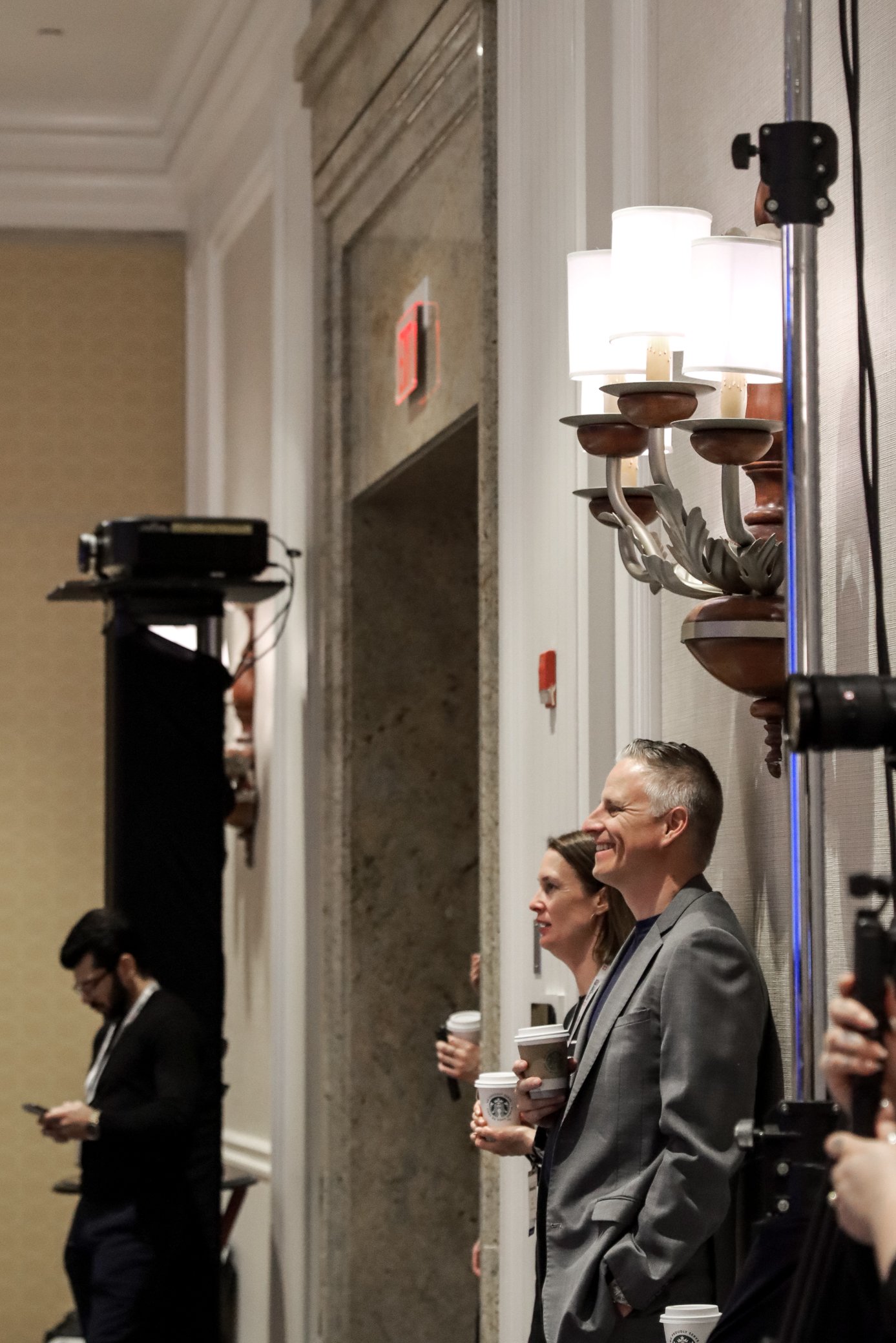 People standing in a line at an indoor event, holding coffee cups and looking ahead, with some engaging with their phones. The setting appears to be a conference or seminar room, with a chandelier on the wall and a speaker or clapperboard setup in th