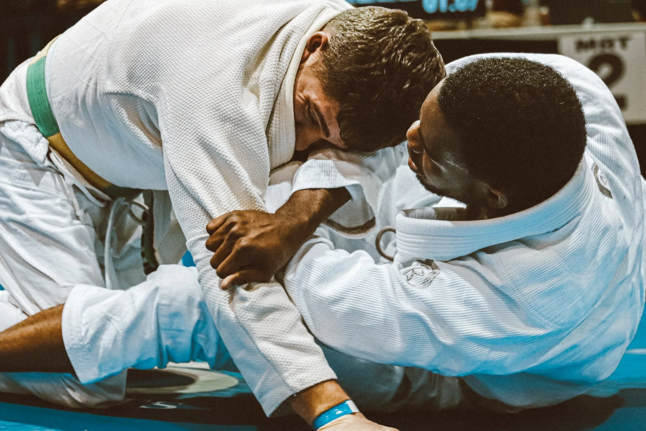 Two male judo competitors engaged in a grappling match on the mat, wearing white judogi with colored belts, one with a green belt and the other with a black belt.