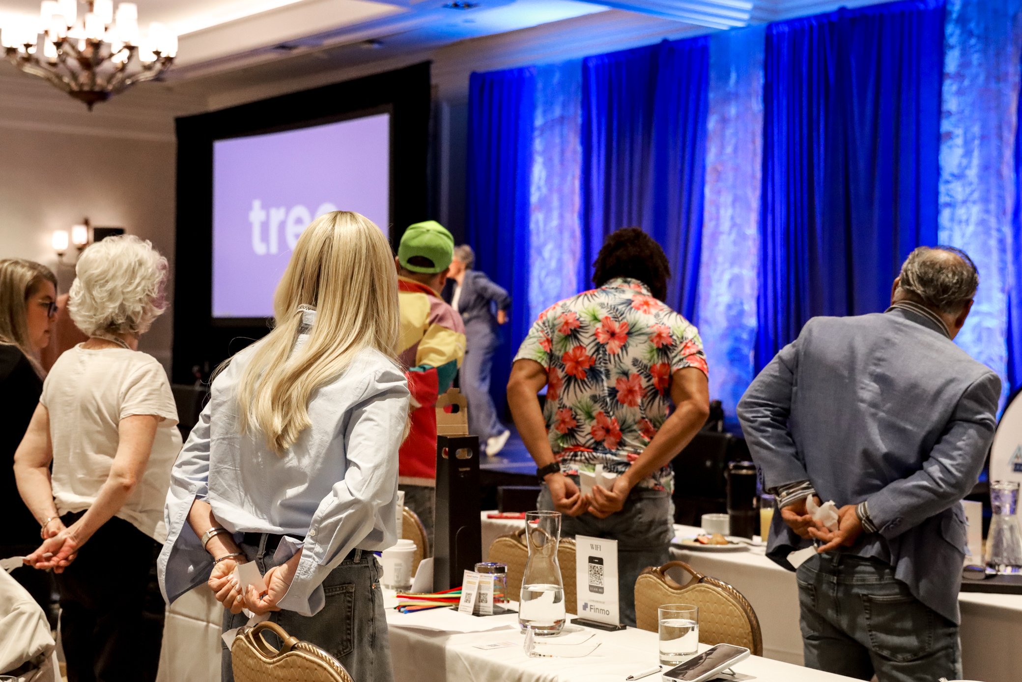 People attending a conference or meeting, standing around tables with drink glasses and informational materials in a decorated hotel conference room with a large blue curtain and a projected screen.
