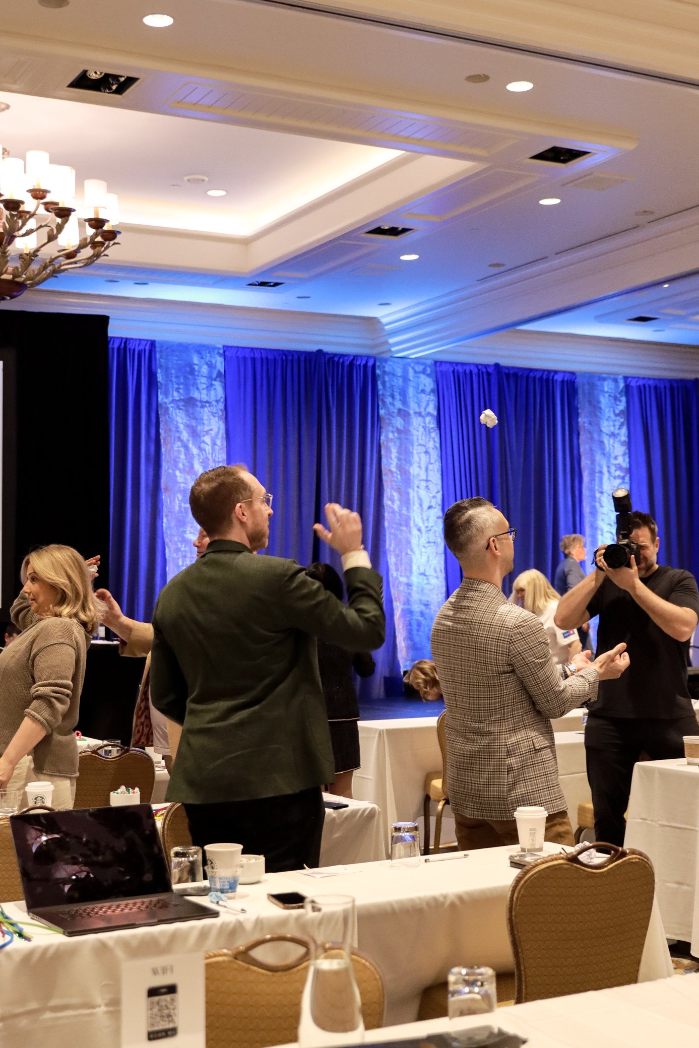 Conference room with people standing, talking, and taking photos. Tables with cups, bottles, and laptops, blue curtains on the stage, chandelier on ceiling, and some chairs.