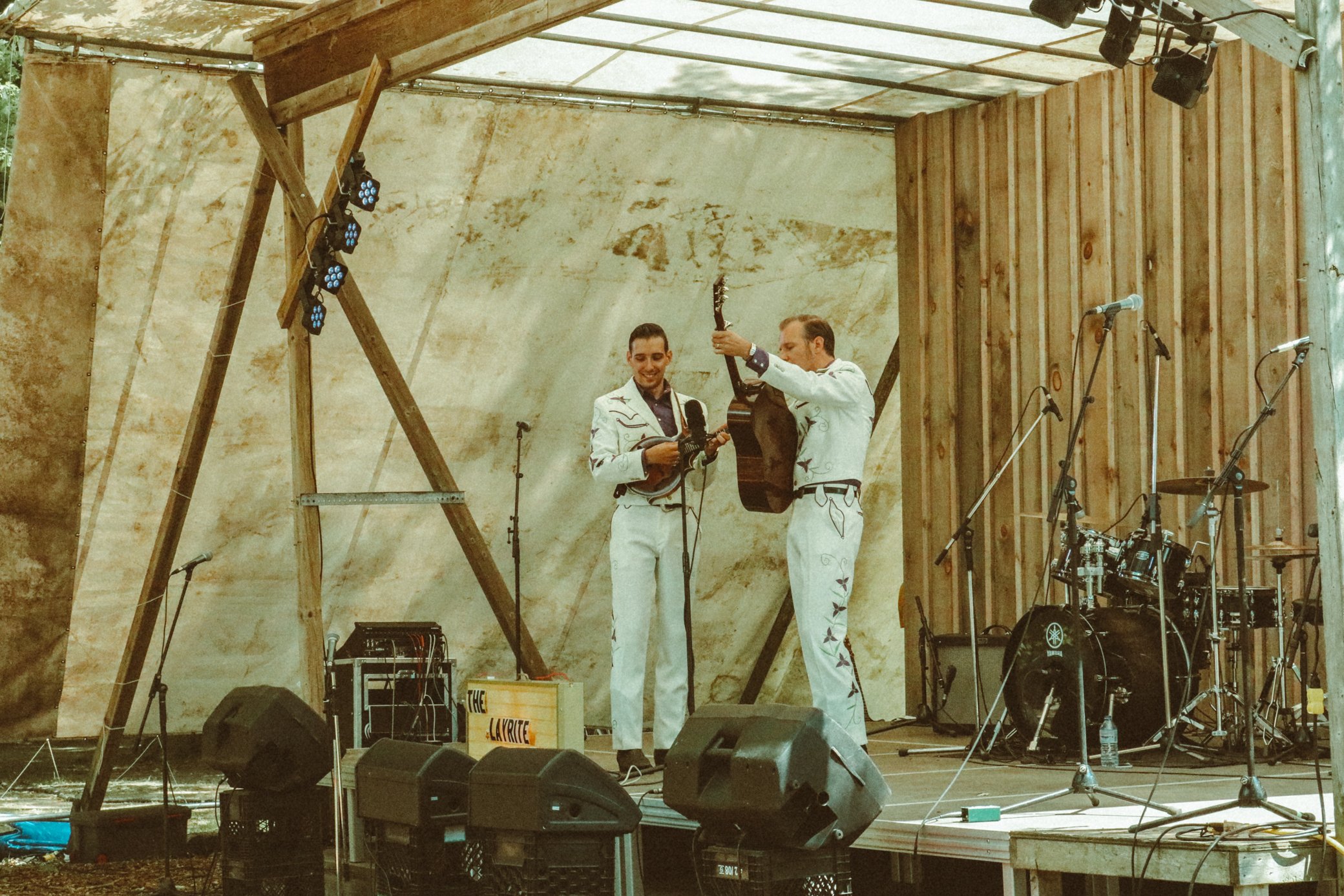 Two men in matching white suits with black embroidered designs on stage, one playing a guitar and the other holding a microphone. The stage is set outdoors with wooden panels, microphones, speakers, and musical instruments, and stage lights hanging f