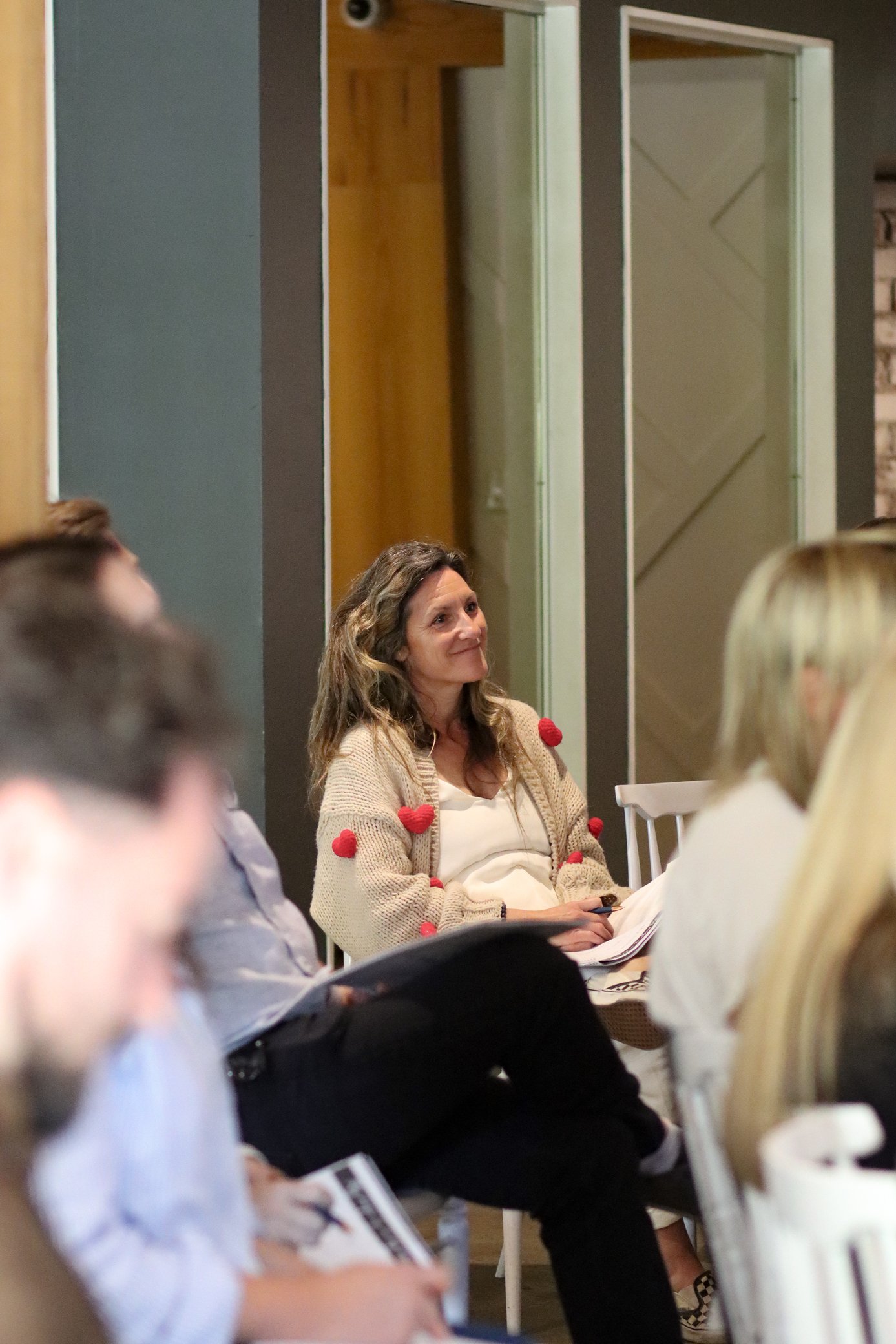 A woman with wavy brown hair sitting at a table during a gathering or meeting, wearing a beige cardigan with red pom-poms, holding and looking at a notebook, with other people partially visible around her.