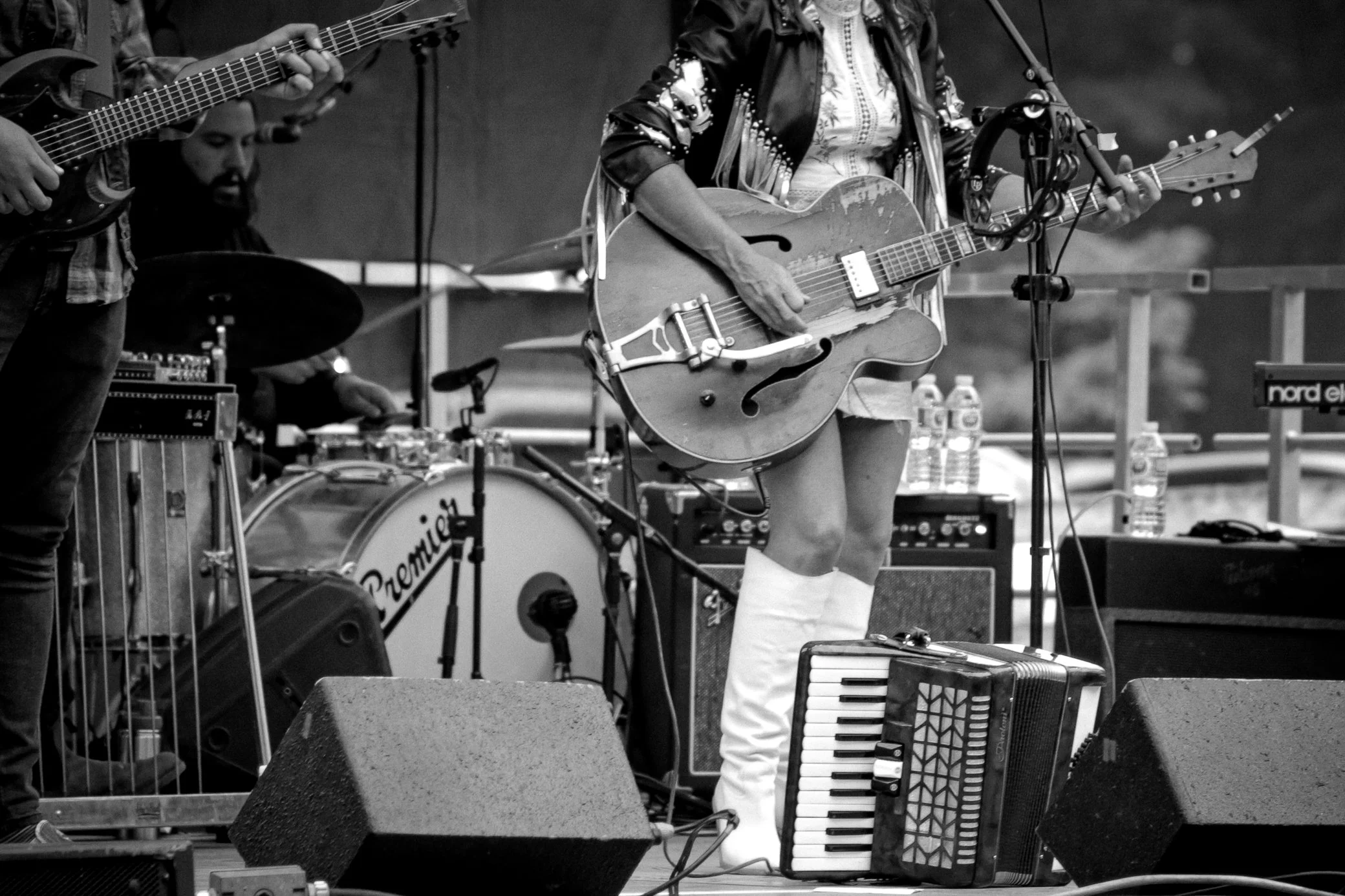 Musicians performing on stage: a person playing an electric guitar, a person playing an acoustic guitar, and a person playing an accordion, with musical equipment around them.