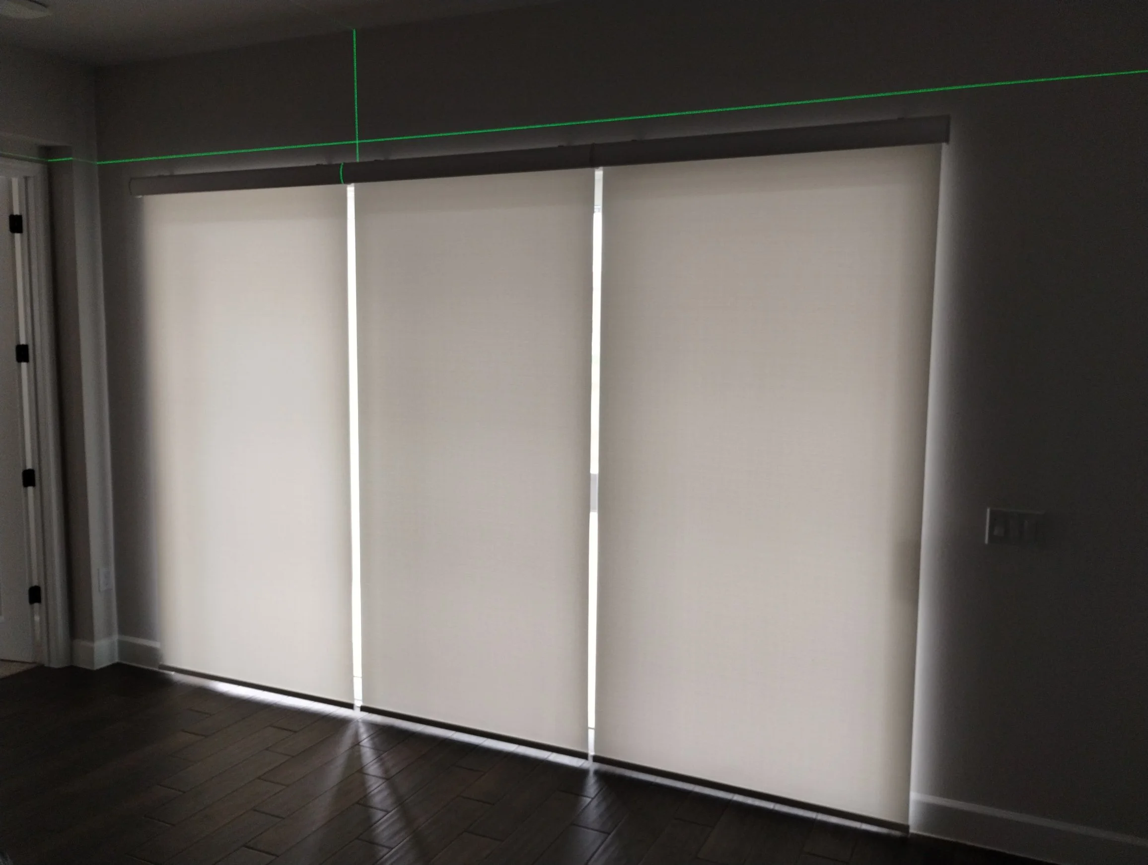 Interior view of a room with a large window covered by vertical blinds, with a glowing green laser line projecting across the ceiling and wall.