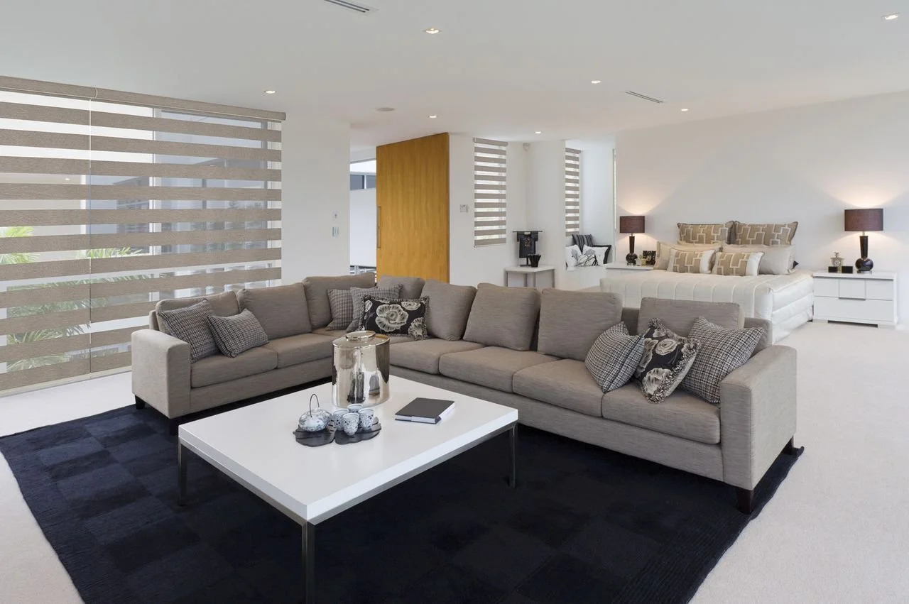 Modern living room with beige sectional sofa, black area rug, white coffee table with decorative items, and a bedroom in the background with a large bed, white bedding, and two dark lamps.