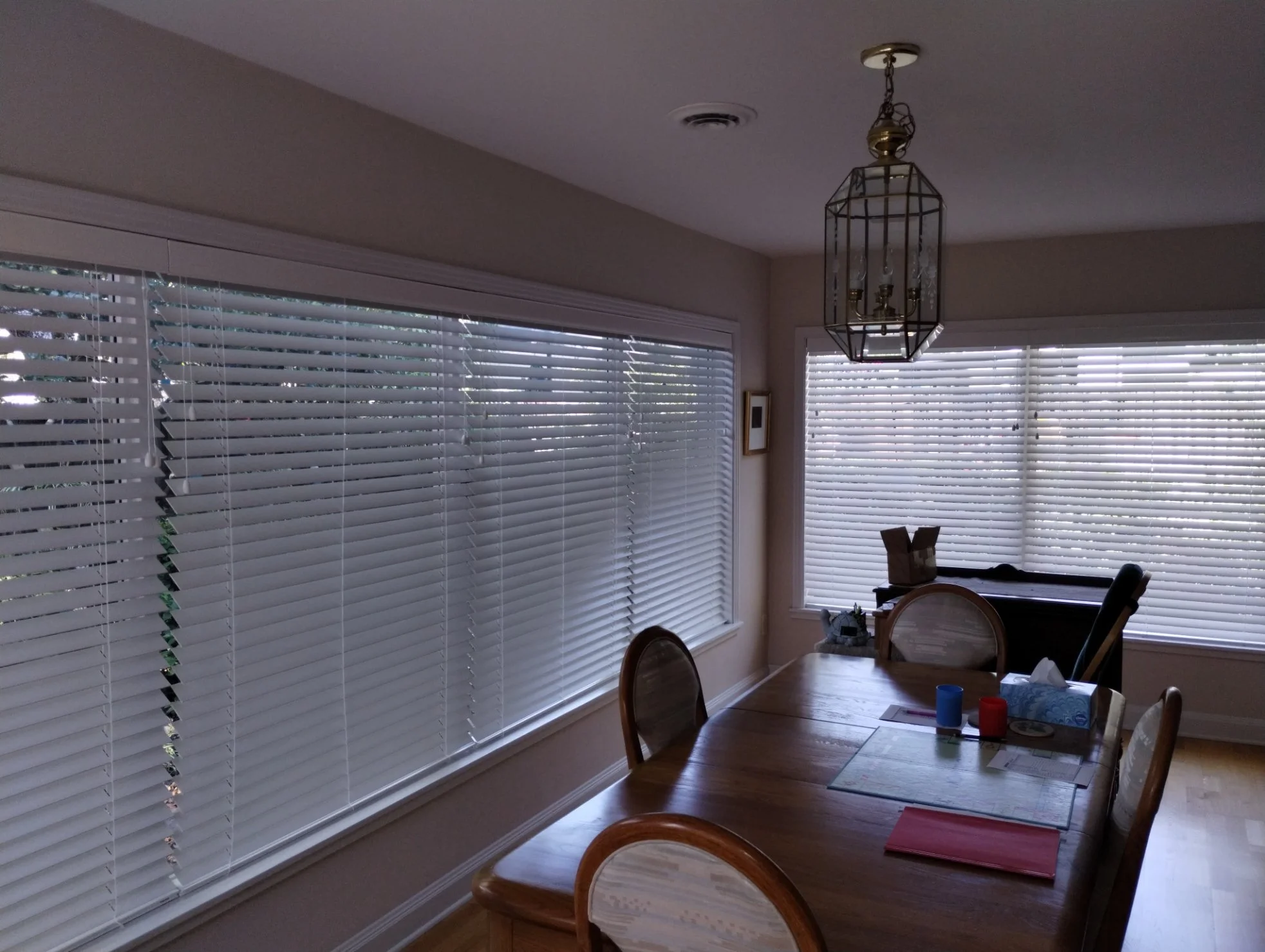 A dining room with hardwood floor and large windows with white horizontal blinds. A wooden dining table with papers, cups, and tissue box, surrounded by wooden chairs, is visible. An ornate chandelier hangs from the ceiling.