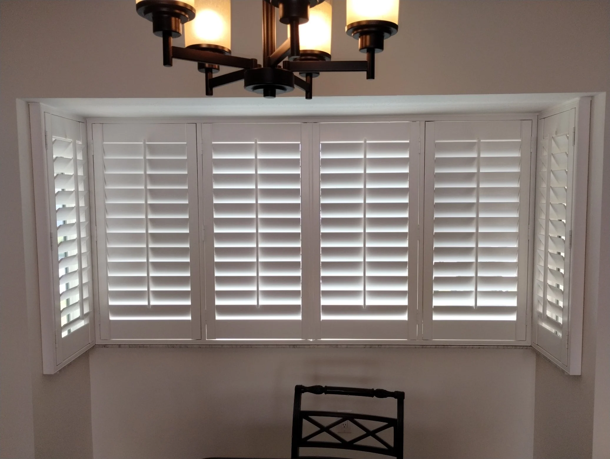A picture of a room with white plantation shutters on a window, a black metal chair in front of the window, and a modern chandelier hanging from the ceiling.
