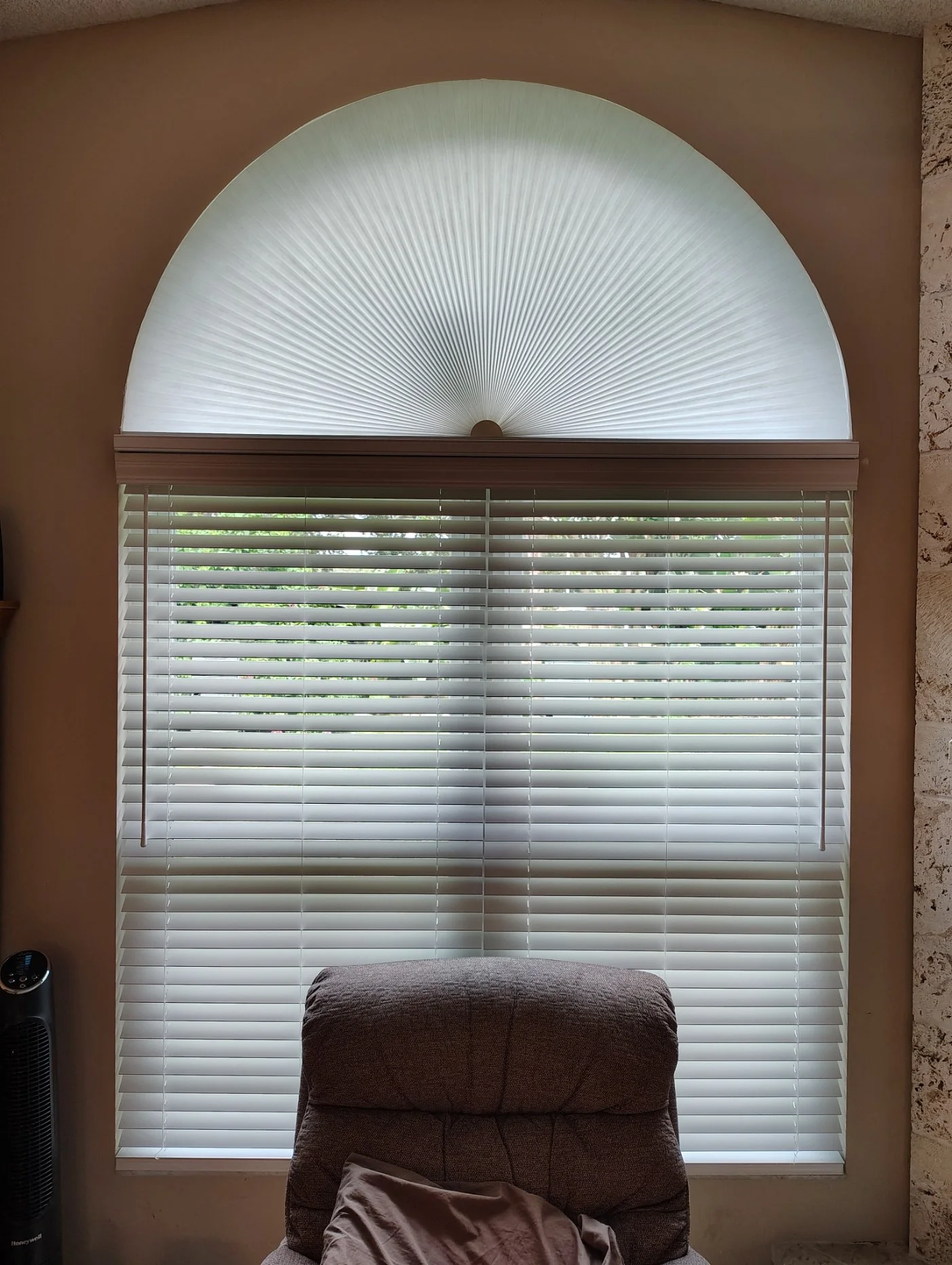 A window with a semi-circular top covered by a light filter, with closed horizontal blinds below. A brown armchair with a blanket or clothing draped over it in front of the window.