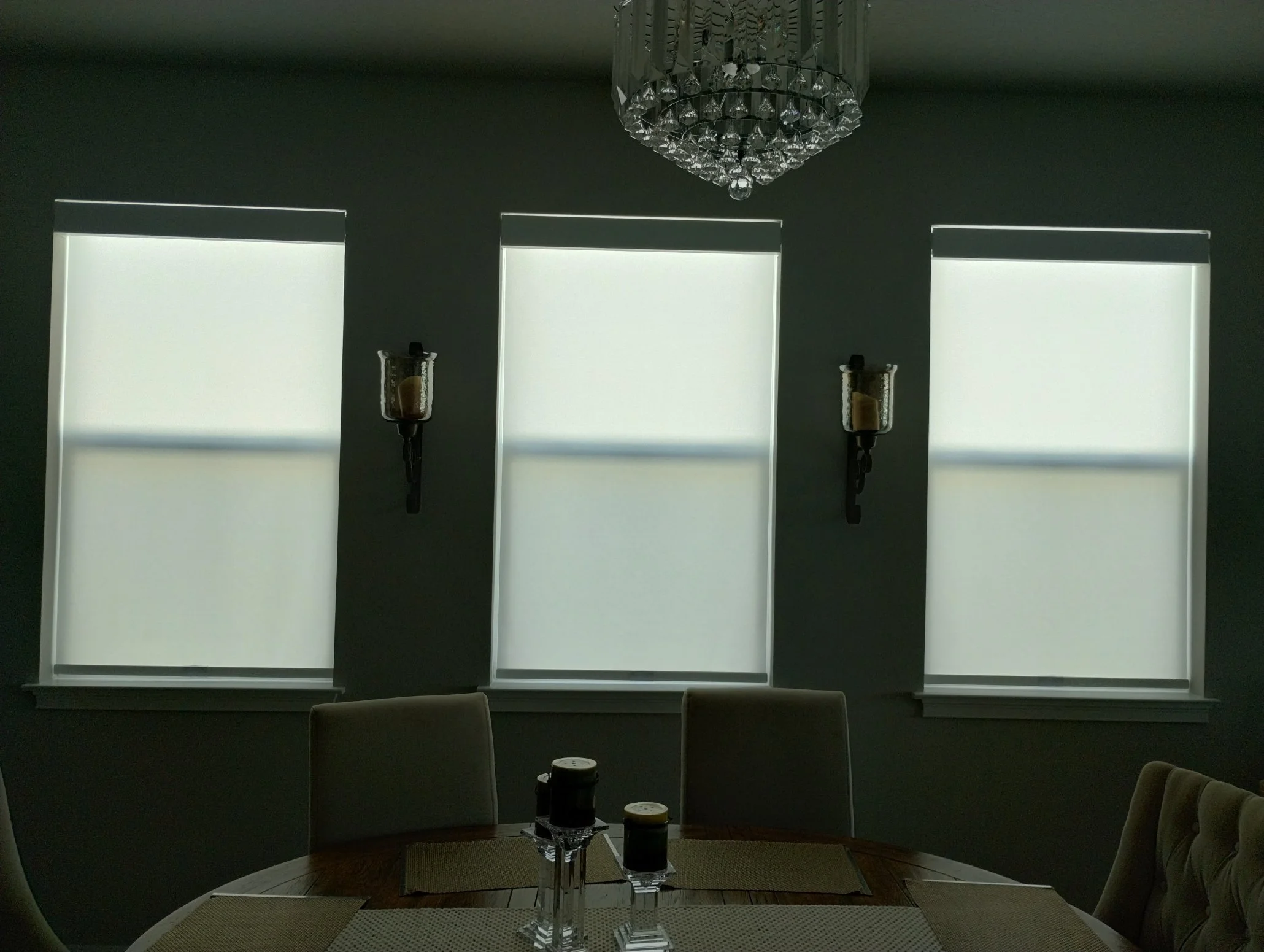 A dining room with three windows covered by white blinds, a chandelier hanging from the ceiling, two wall-mounted candle holders with candles, and a table with three coffee cups.
