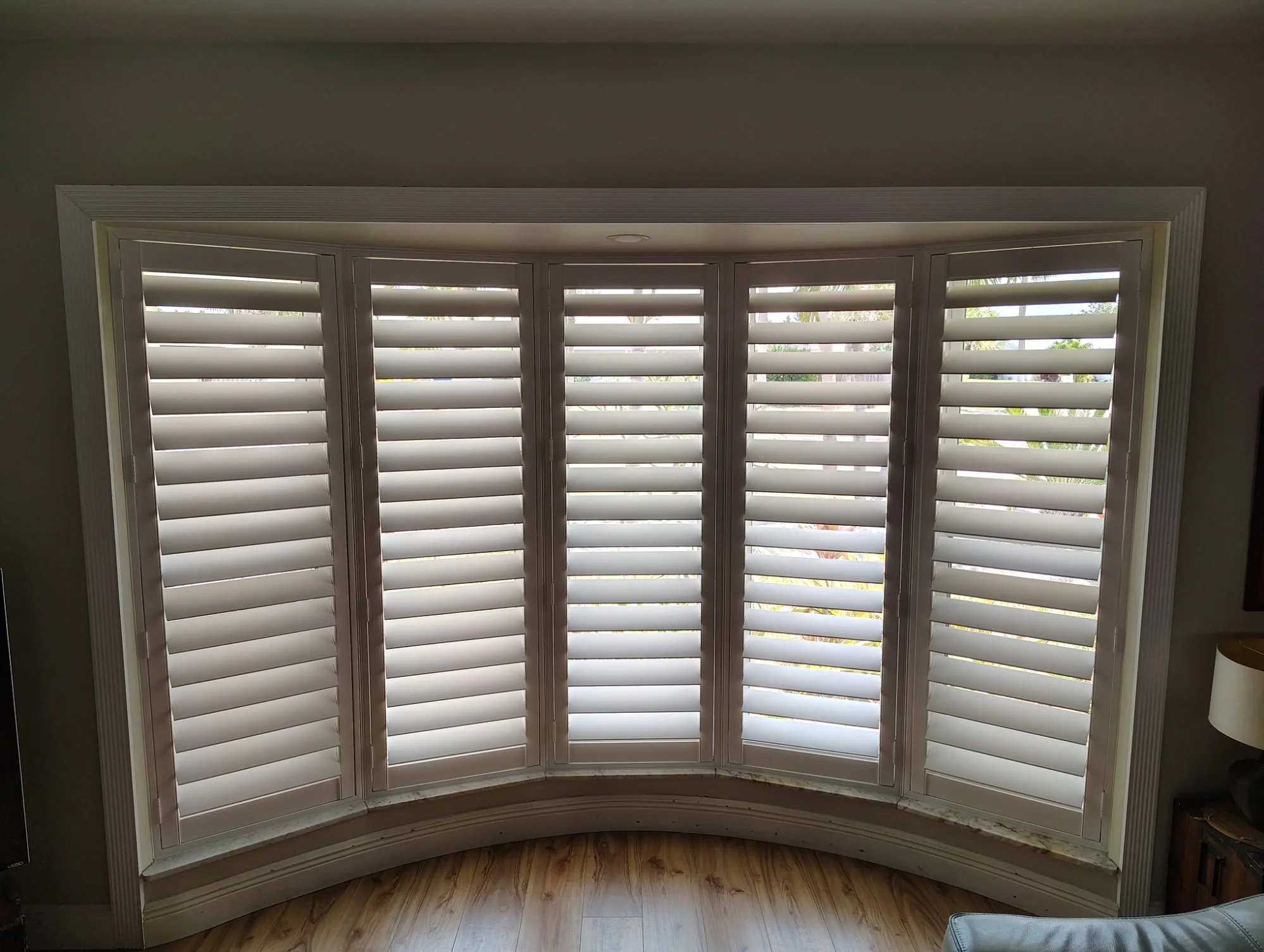 Curved window with white plantation shutters covering all window panels, located in a room with hardwood flooring and a neutral wall color.
