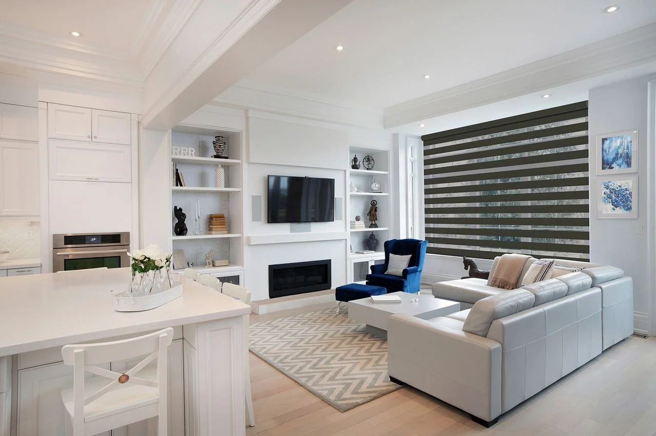 Bright, modern living room with white walls and furniture, blue accent chair, striped blackout window shades, TV above fireplace, built-in shelves with decor, and artwork on the walls.