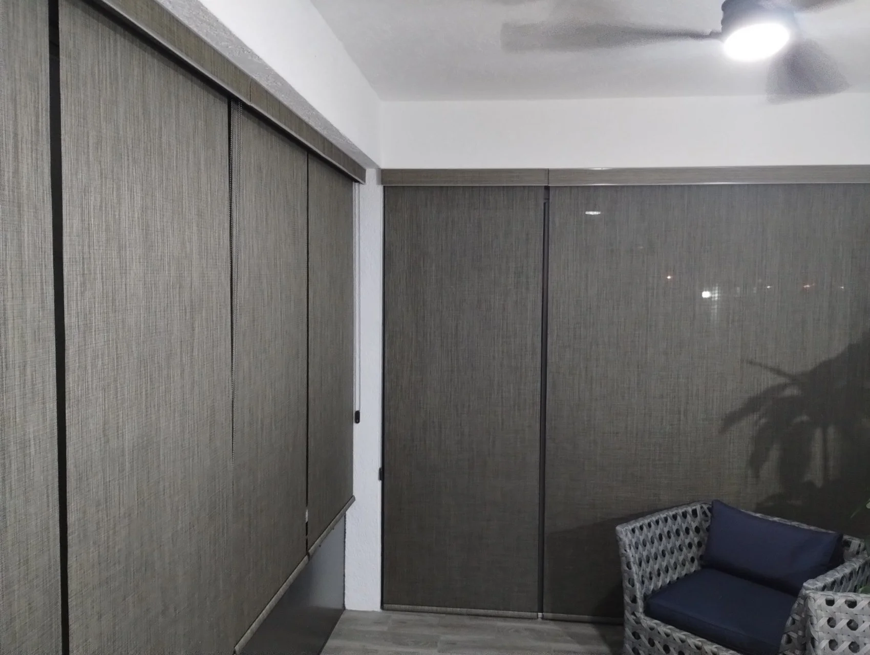 Balcony or patio area with sliding glass doors and woven patio furniture with cushions, illuminated by a ceiling light.