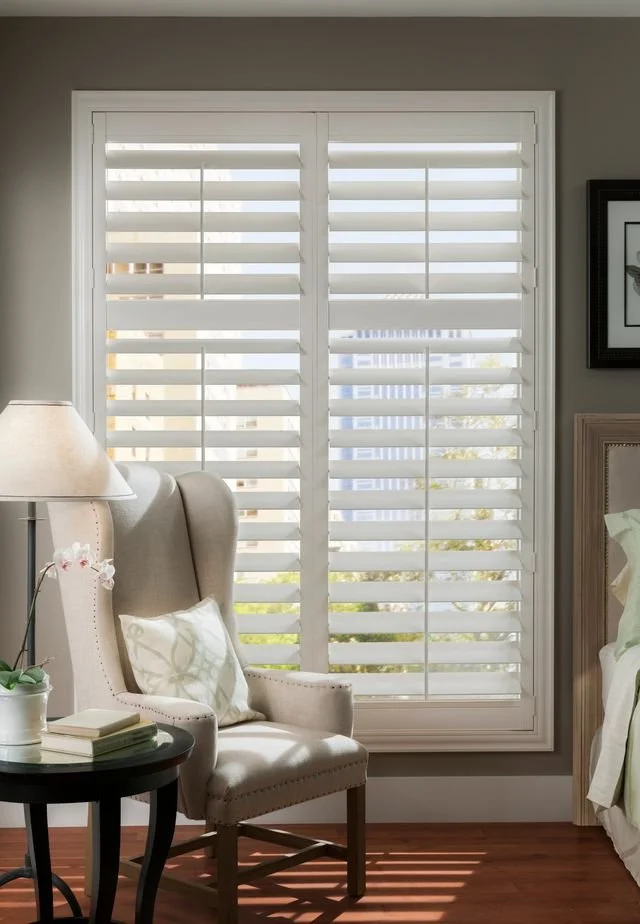 Living room with a large window featuring white plantation shutters, an upholstered armchair with a pillow, a side table with books and a potted plant, and a lamp. Part of a bed and framed artwork are visible.