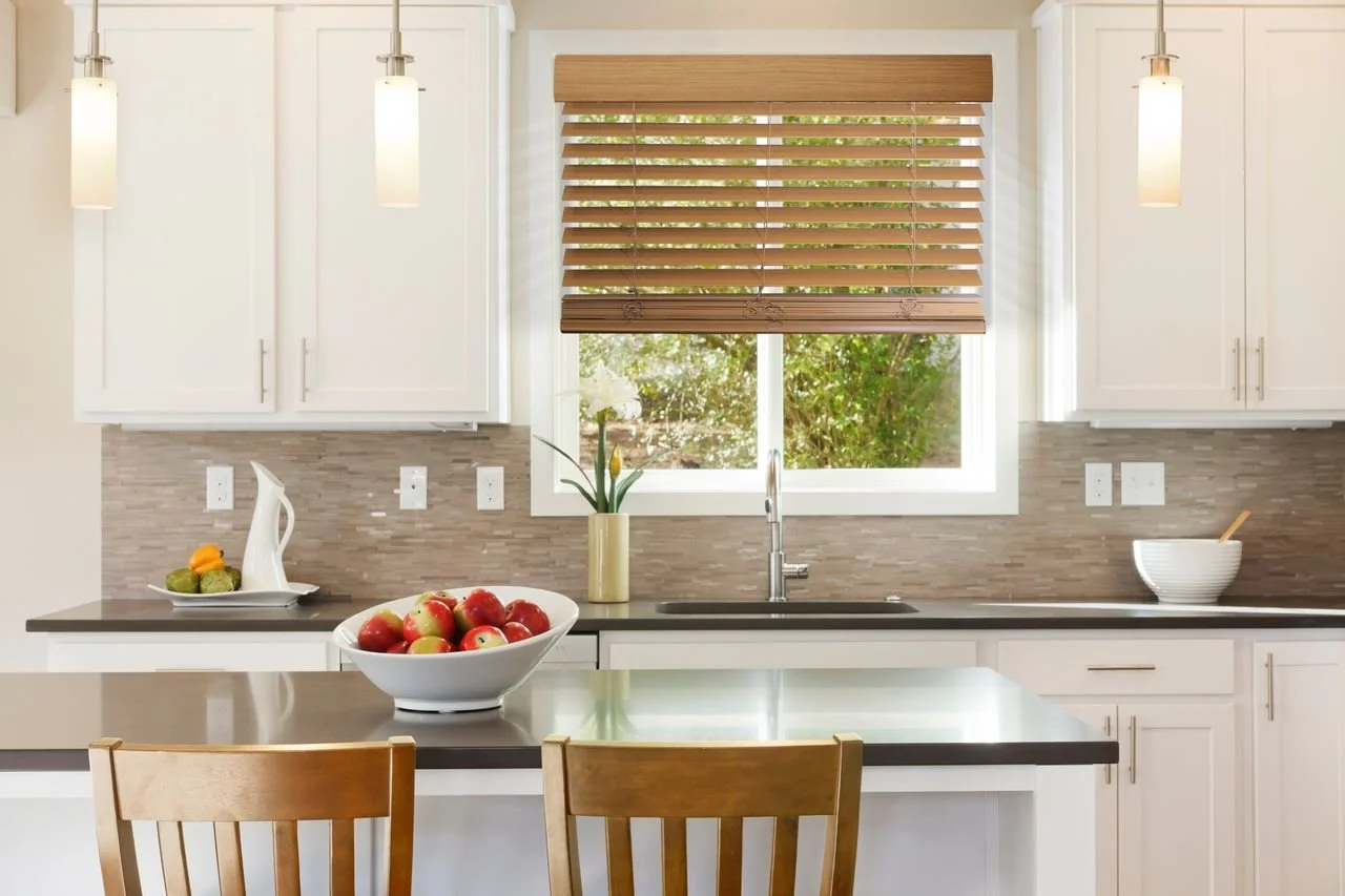 Modern kitchen with white cabinets, a window with blinds, pendant lights, a bowl of apples on the island, a vase with flowers, and a bowl with a spoon.