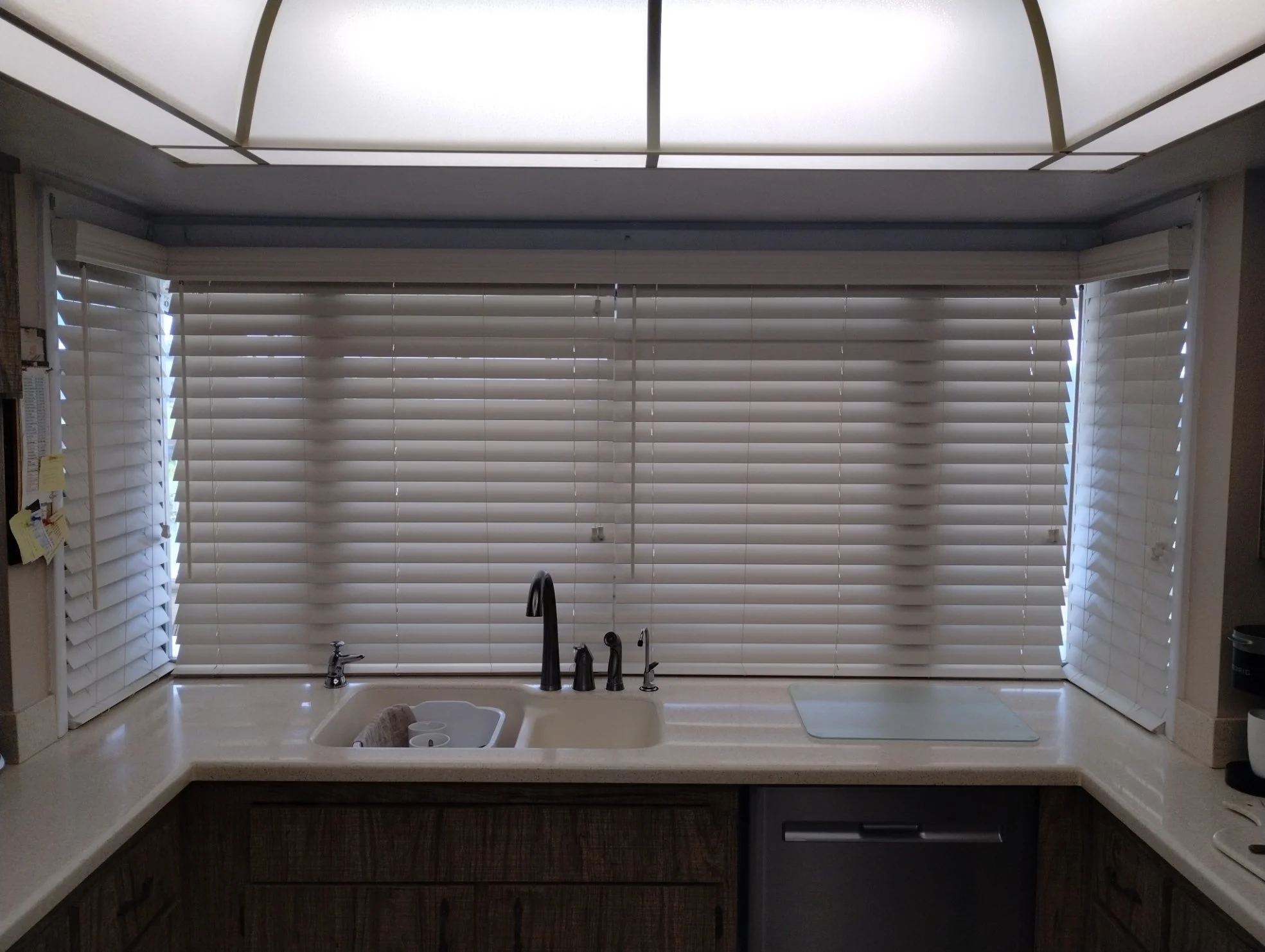Kitchen window with white blinds and ceiling light, sink with dark faucet, countertop with a small white tray, and kitchen cabinets below.