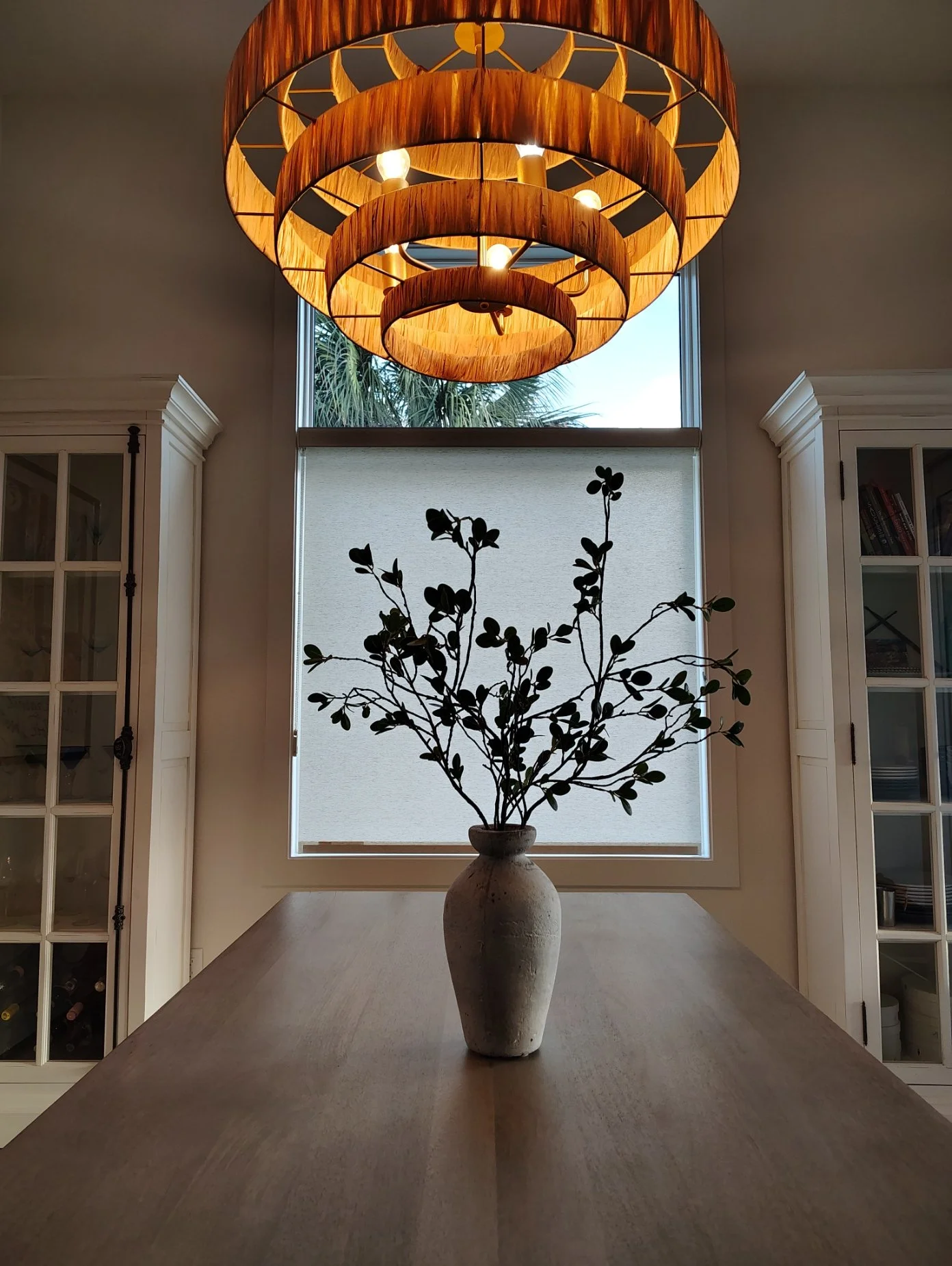 A ceramic vase with leafy branches on a wooden table in front of a large window and a circular wooden chandelier hanging from the ceiling.
