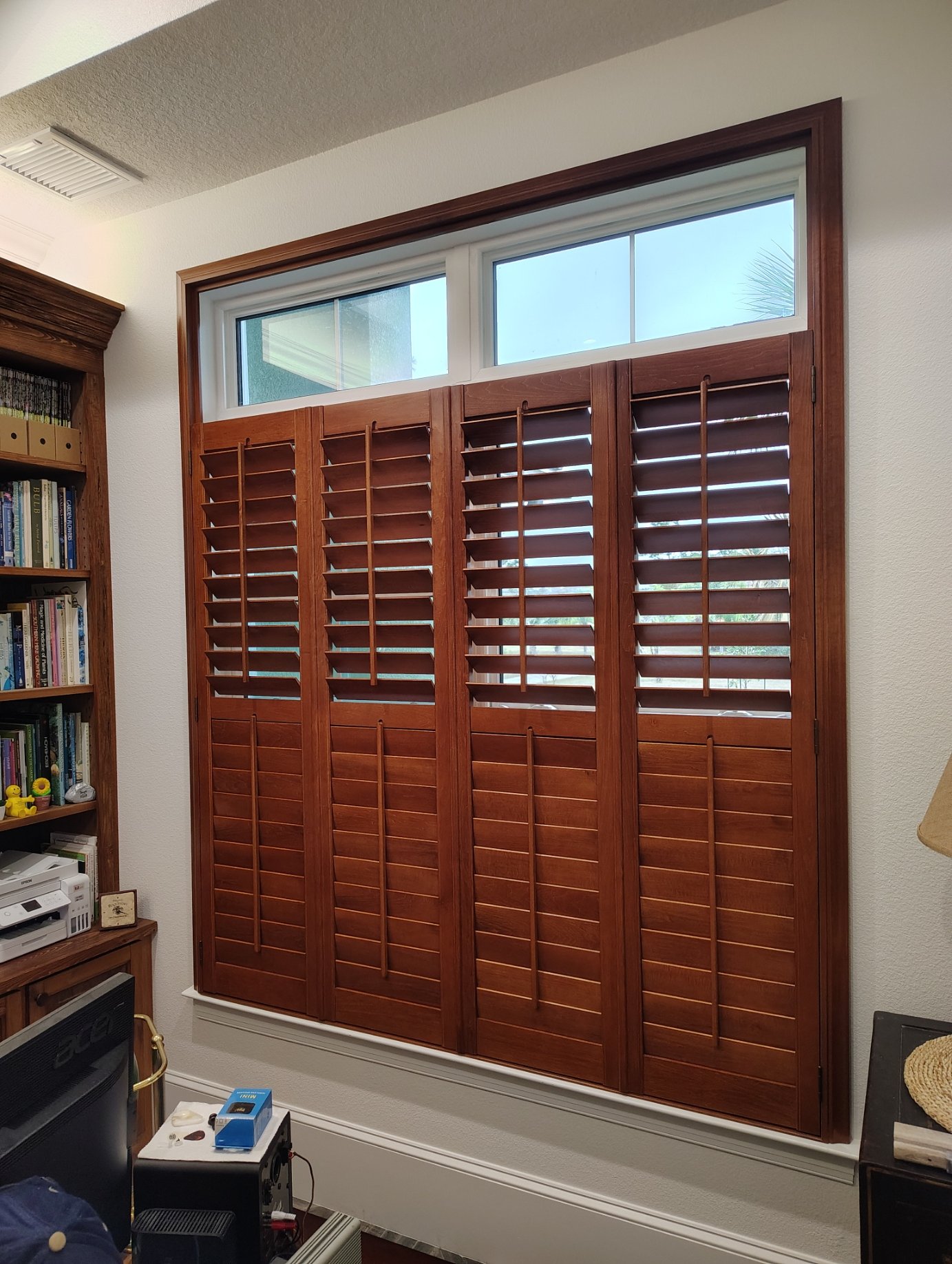 Wooden plantation shutters on a window in a room with bookshelves and office equipment nearby.