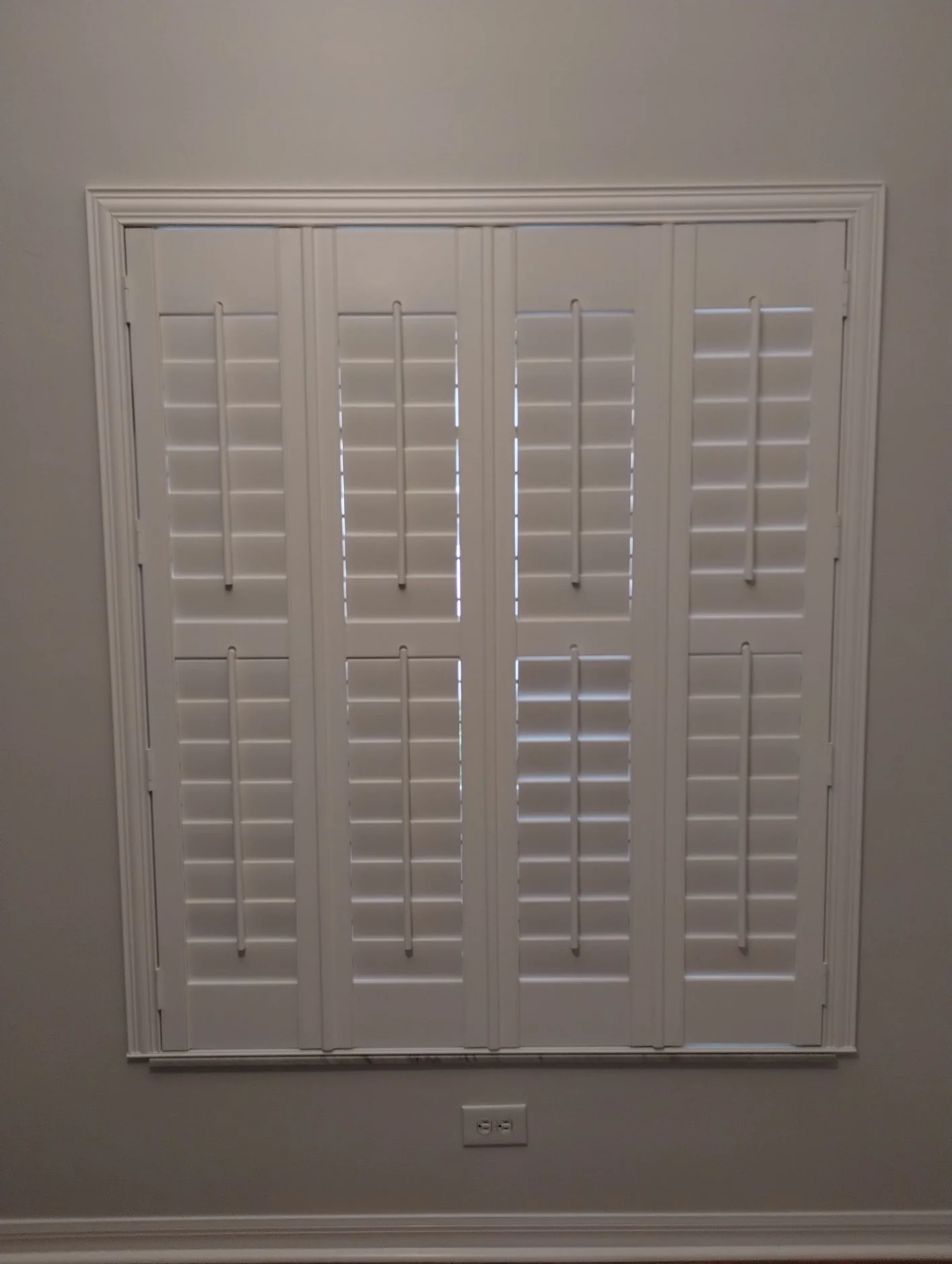 White plantation shutters covering a window on a gray wall, with a power outlet at the bottom.
