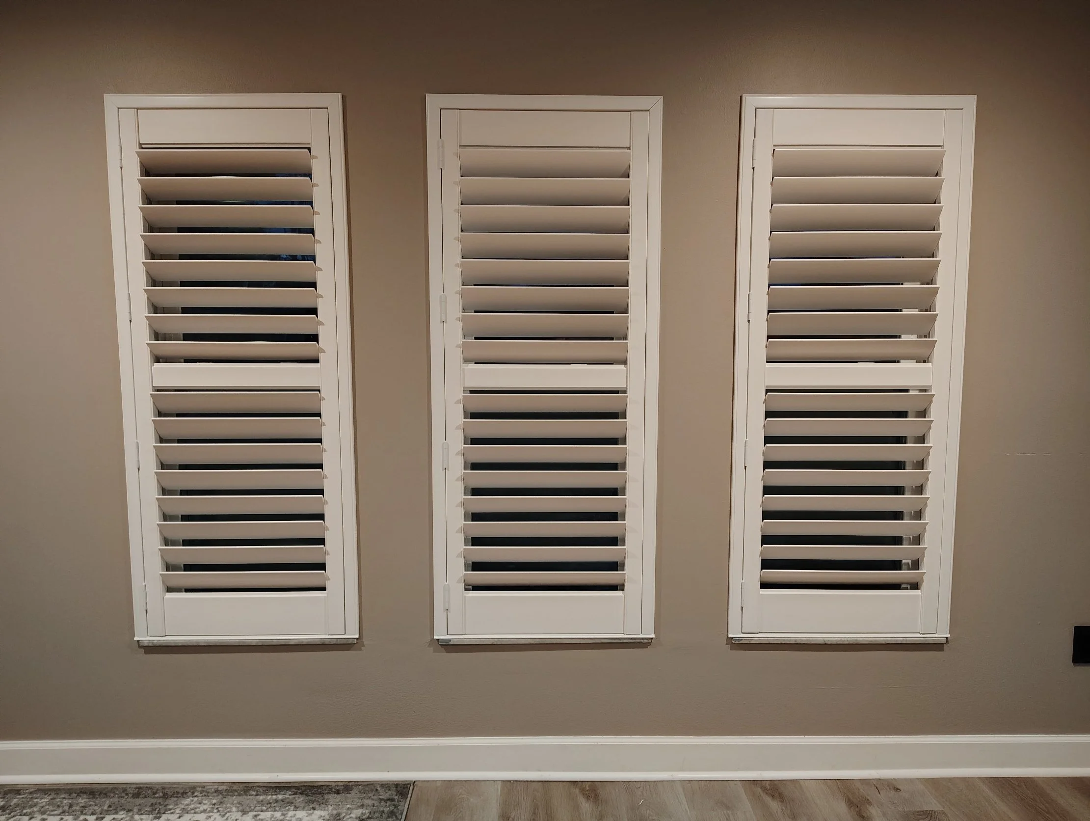 Three white plantation shutters mounted on a beige wall, with a glimpse of a carpeted floor at the bottom.