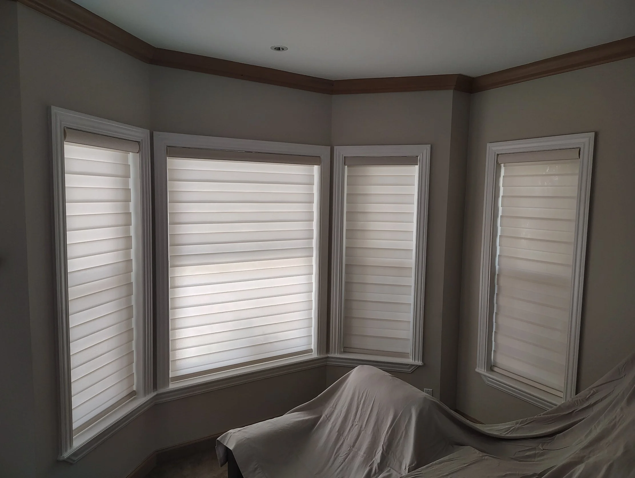 Room with three large windows fitted with beige blinds, crown molding along the ceiling, and a table covered with a white sheet.