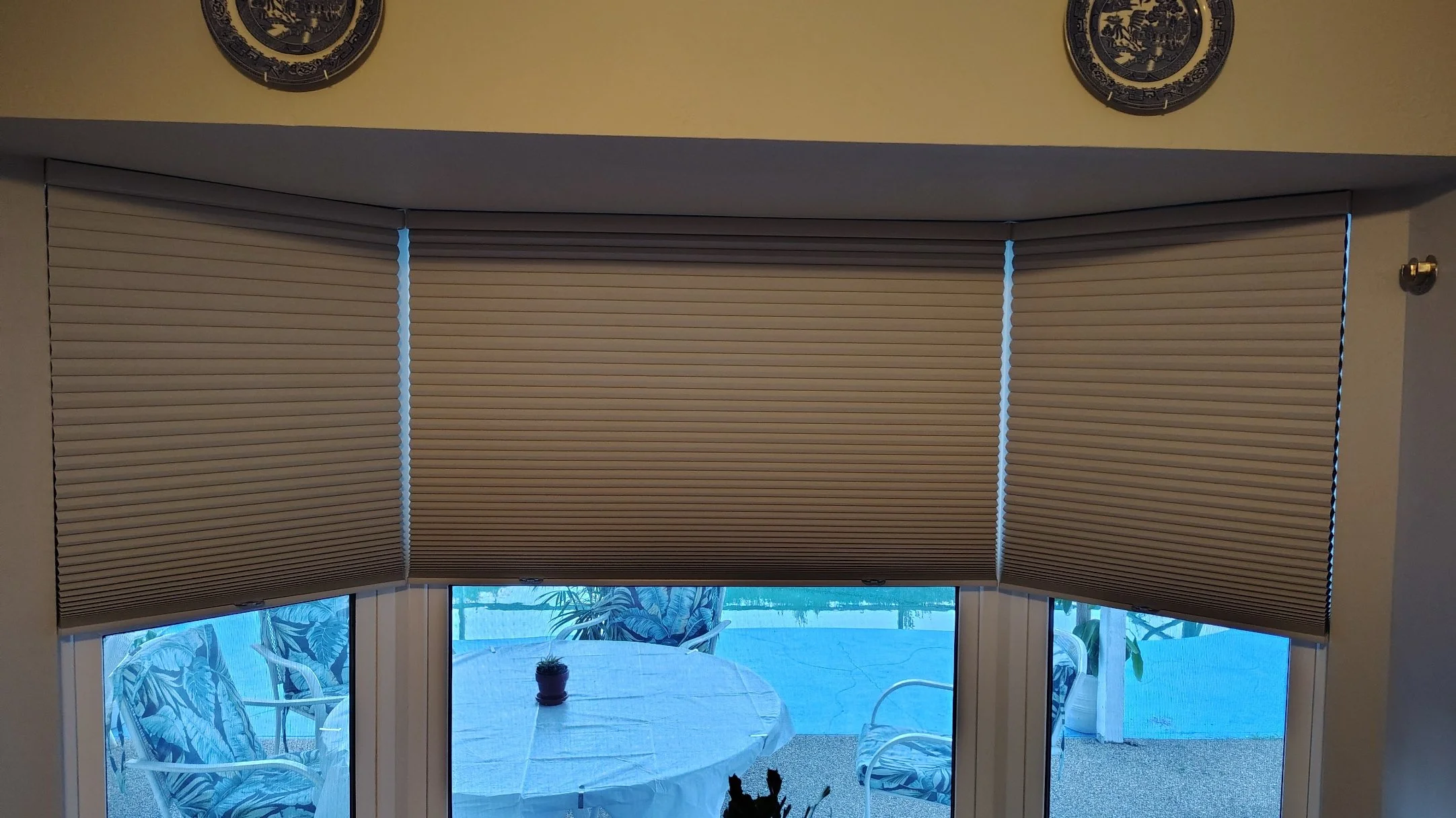 Window with closed beige blinds, a sliding door below with a view of a patio table and chairs, potted plant, and ocean or lake scenery in the background.