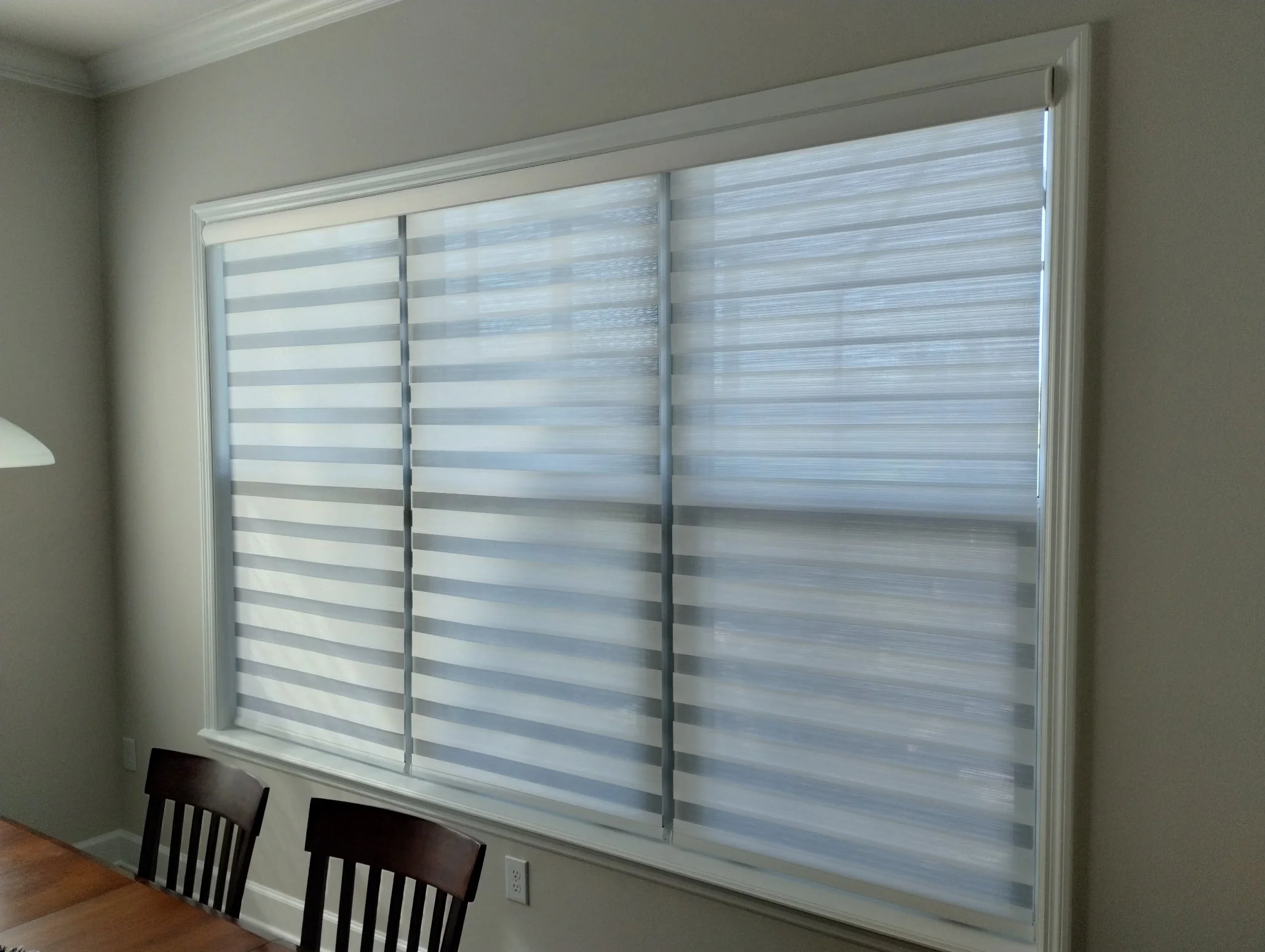 Interior view of a room with a large window covered by zebra blinds. There are dark wood chairs at a dining table at the bottom of the image. The walls are painted in a neutral color, and the room has crown molding at the ceiling.