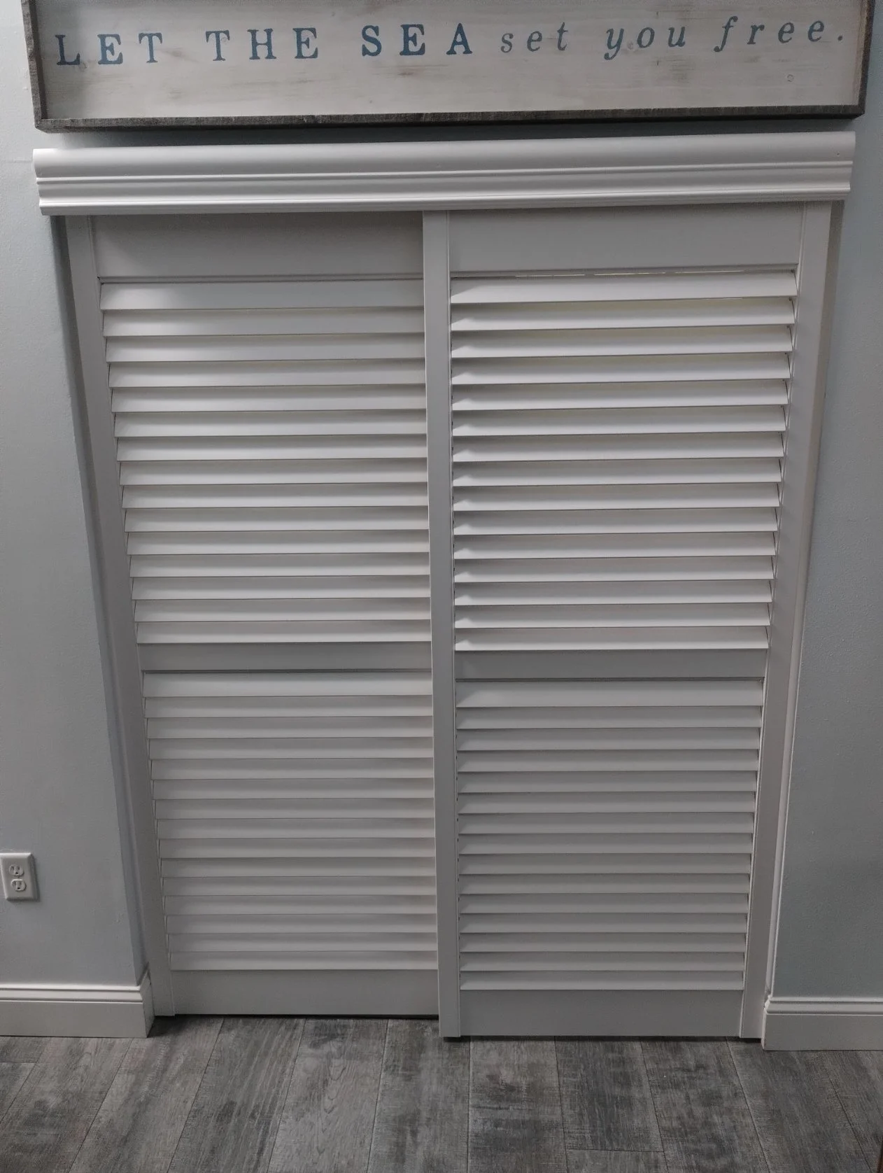 A white louvered closet door with a horizontal trim at the top. Above the door, a sign with the phrase "Let the sea set you free" is visible. To the left of the door, an electrical outlet is slightly visible on a gray wall, and the floor has a wood-l