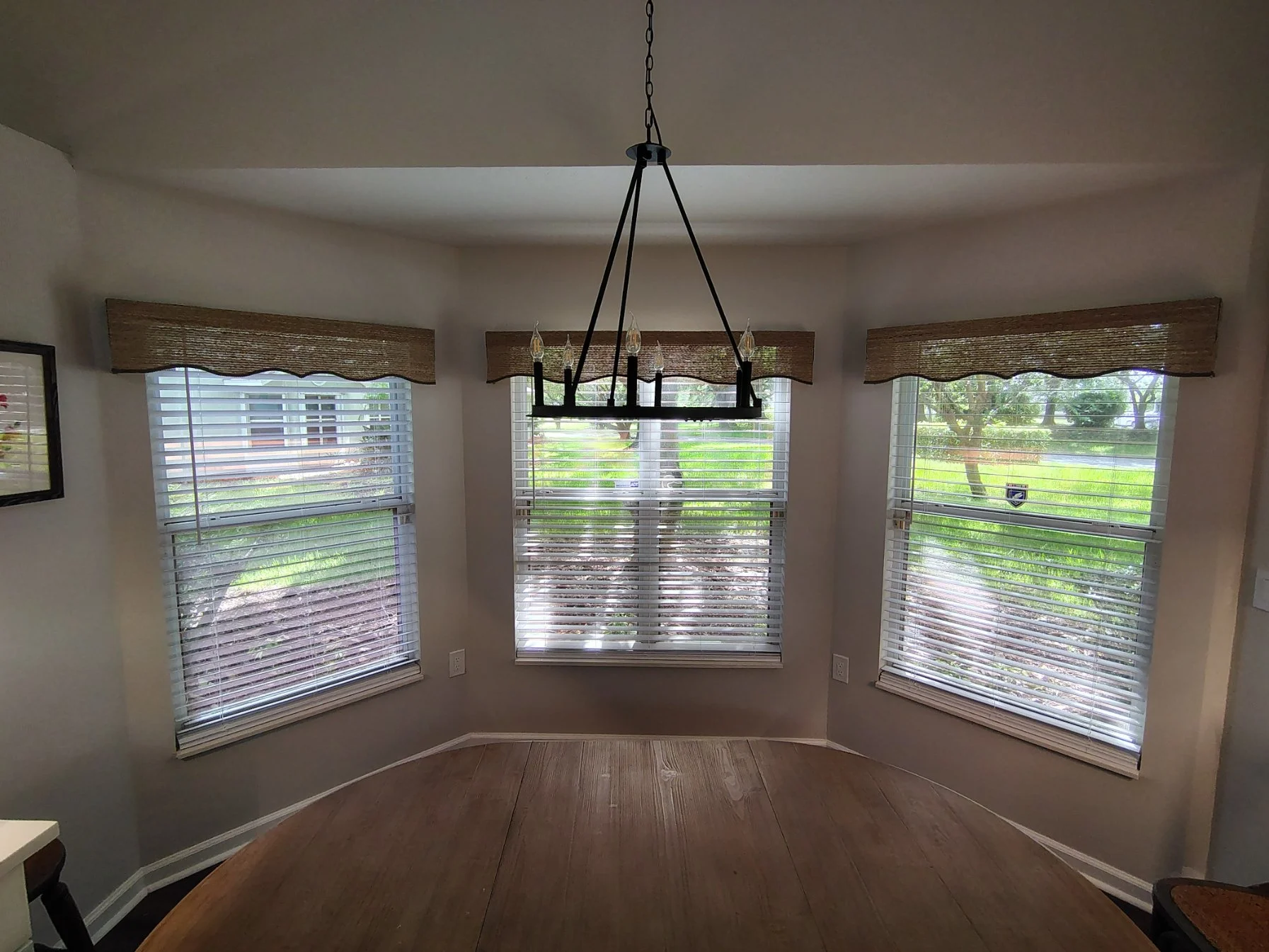 A bright corner of a room with three large windows with white blinds and brown woven valances, a black chandelier hanging from the ceiling, and a wooden dining table in the foreground.