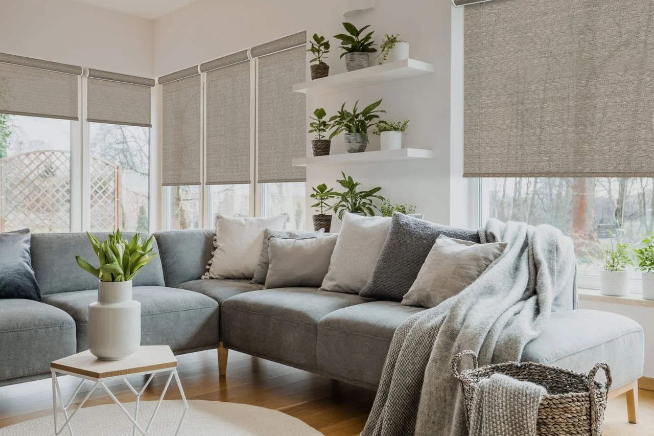 A bright living room with a large gray sectional sofa, beige and gray pillows, and a throw blanket. There are multiple potted plants on wall-mounted shelves and on the windowsill. A small white side table with a potted plant is in front of the sofa, and a laundry basket with a knitted blanket is nearby. The room has large windows with beige shades, allowing natural light to fill the space.