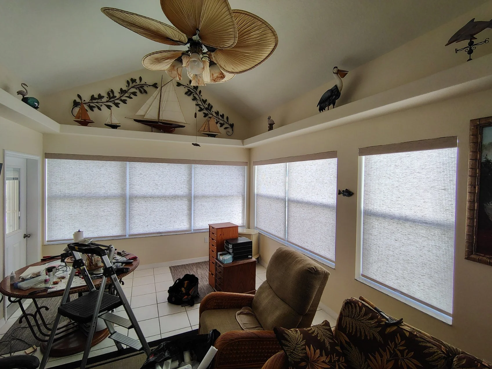 Interior of a room with large covered windows, a ceiling fan with leaf-shaped blades, a small table with various items, a recliner chair, and a sofa with floral upholstery.