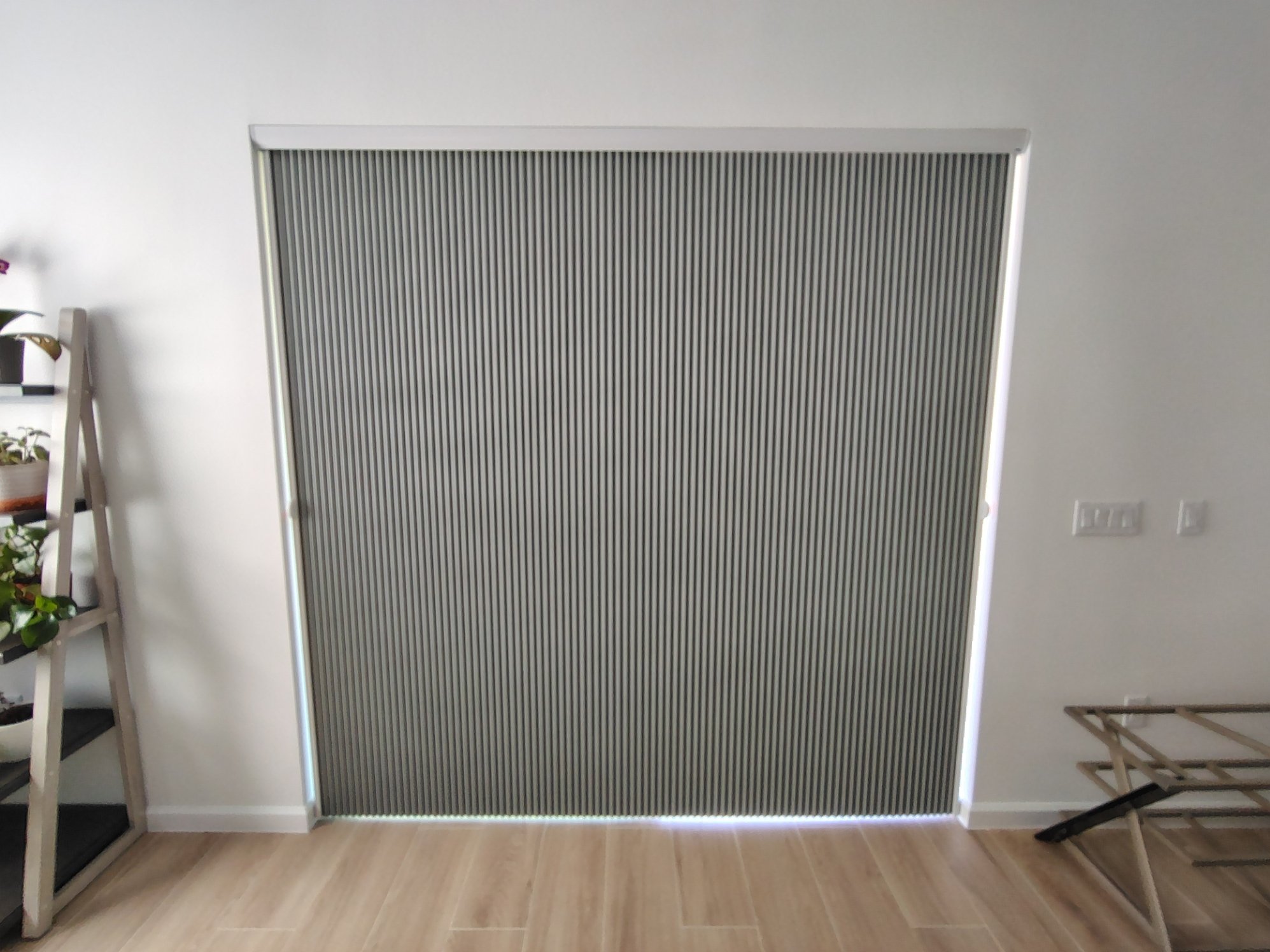 An interior room with a sliding door with a vertical striped pattern, light-colored walls, and wood flooring. To the left, a small ladder shelf holds potted plants, and to the right, there is an unfolded easel or stand.
