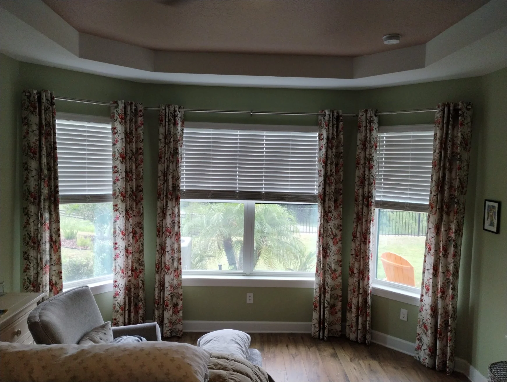 A dining room with large bay windows dressed in floral curtains and white blinds, overlooking a backyard with a patio, chairs, trees, and grass.