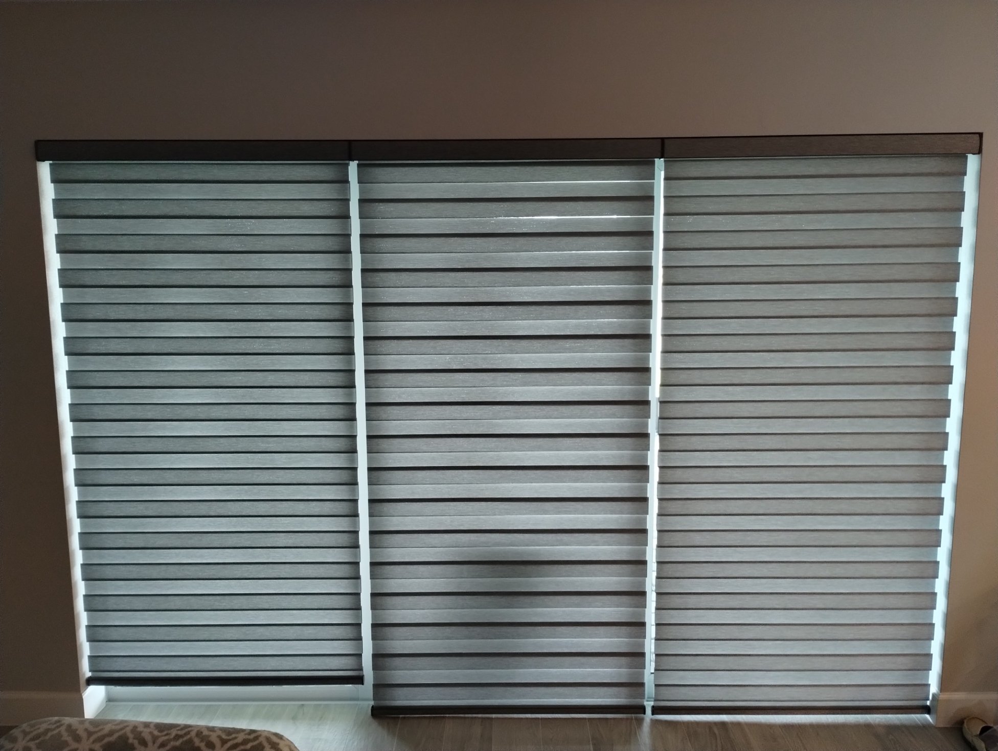 Three window coverings with horizontal slats, two are dark gray and one is a lighter shade of gray, in front of a beige wall.