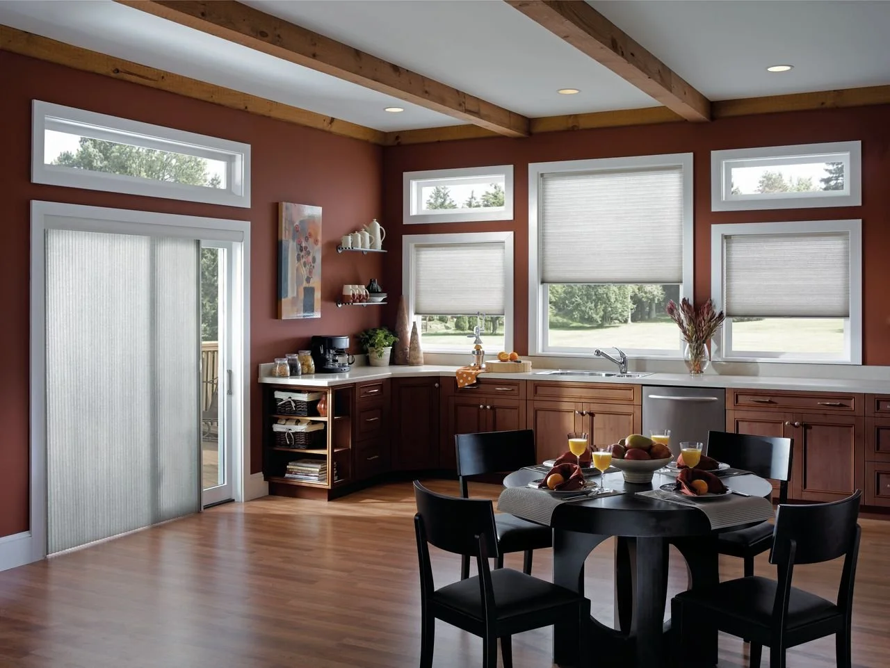 A kitchen with wooden cabinetry, four windows with blinds, a round dining table set with fruit and drinks, and a sliding door leading outside. The room has wooden floors and exposed ceiling beams.
