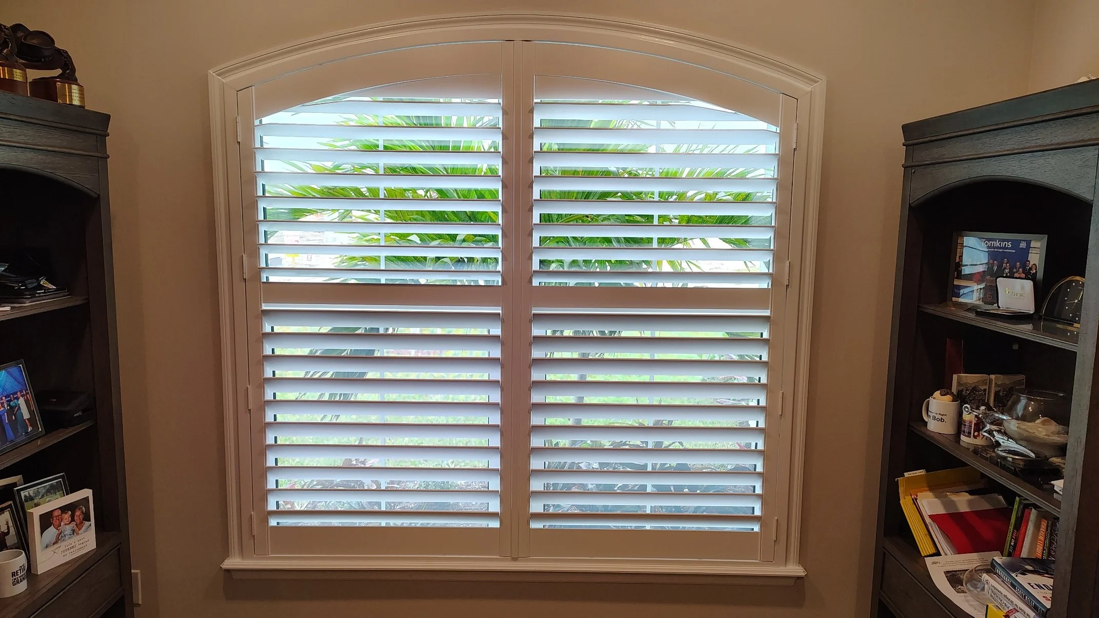 A large arched window with white plantation shutters, partially open, revealing green palm leaves outside. Two dark wood bookshelves flank the window, filled with books, framed photos, and personal items.