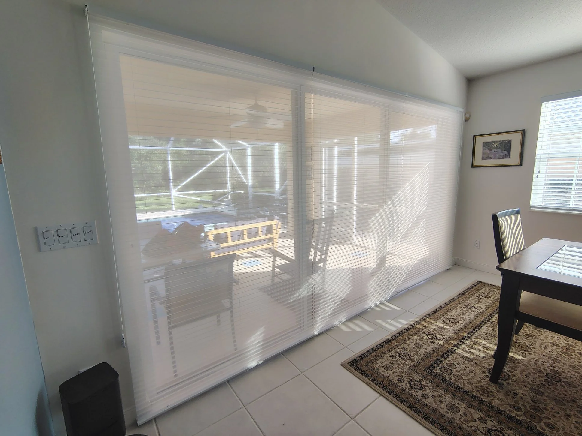 A room with a large sliding window covered by partially open white blinds. There is a patterned rug on the white tiled floor, a framed picture on the wall, a wooden dining table, and a black chair. An outside patio and a ceiling fan are visible throu