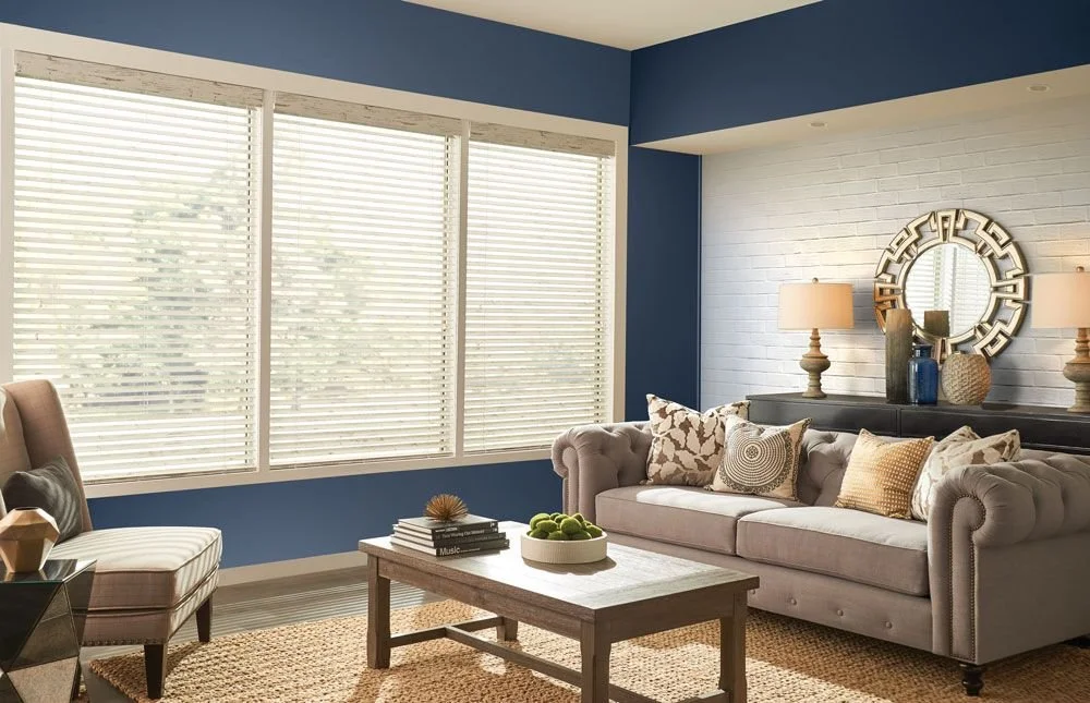 Living room with a beige couch and armchair, wooden coffee table with decorative items, white brick wall with a mirror, and blue walls beside large windows with blinds.