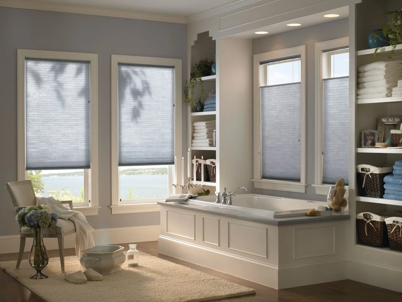Bright bathroom with three windows, a built-in bathtub, shelves with towels and decorative items, a chair with flowers, and a view of water outside.