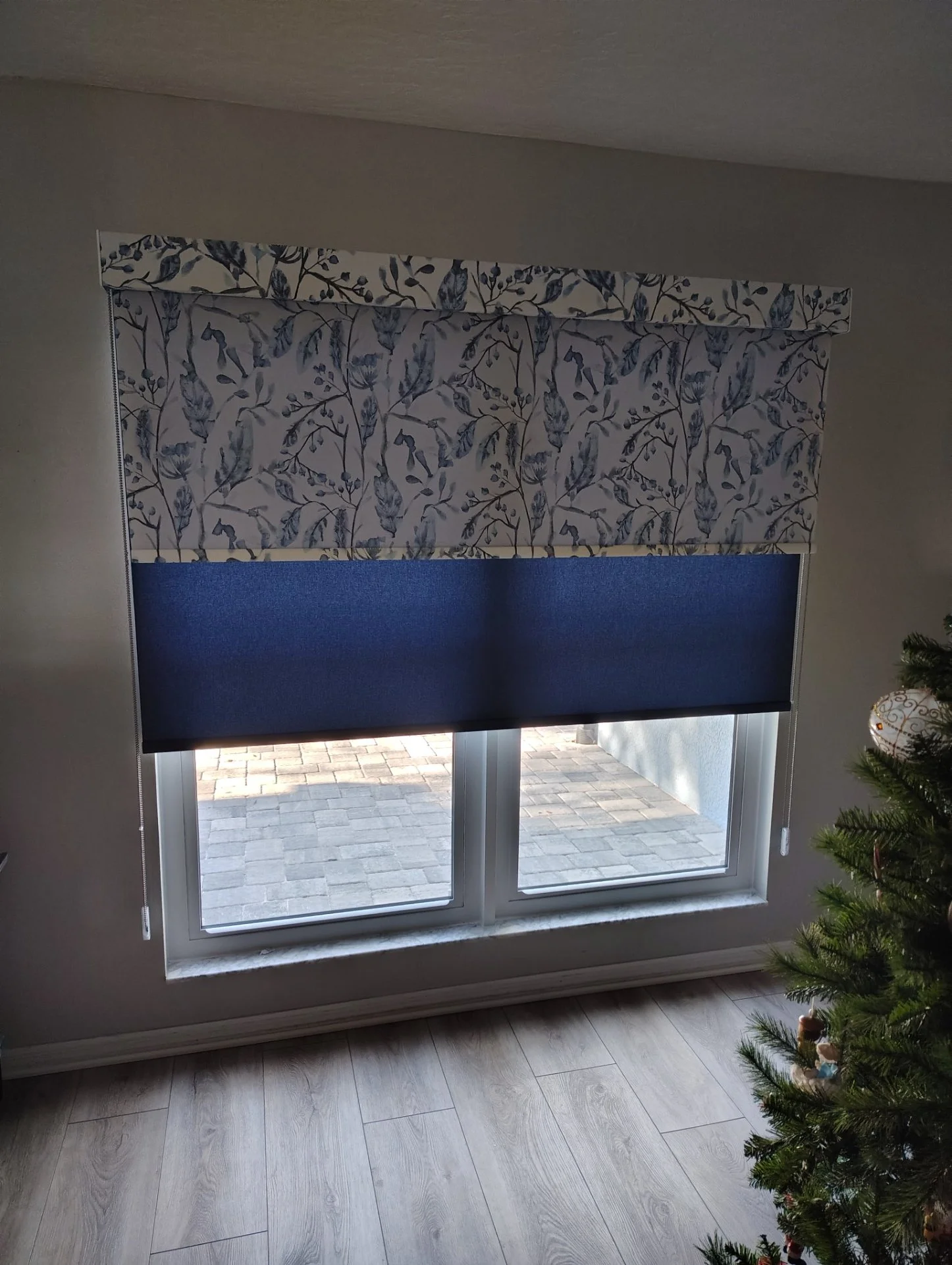 A window with a floral-patterned valance and a blue roller shade, showing a brick patio outside. Part of a Christmas tree with ornaments is visible on the right.