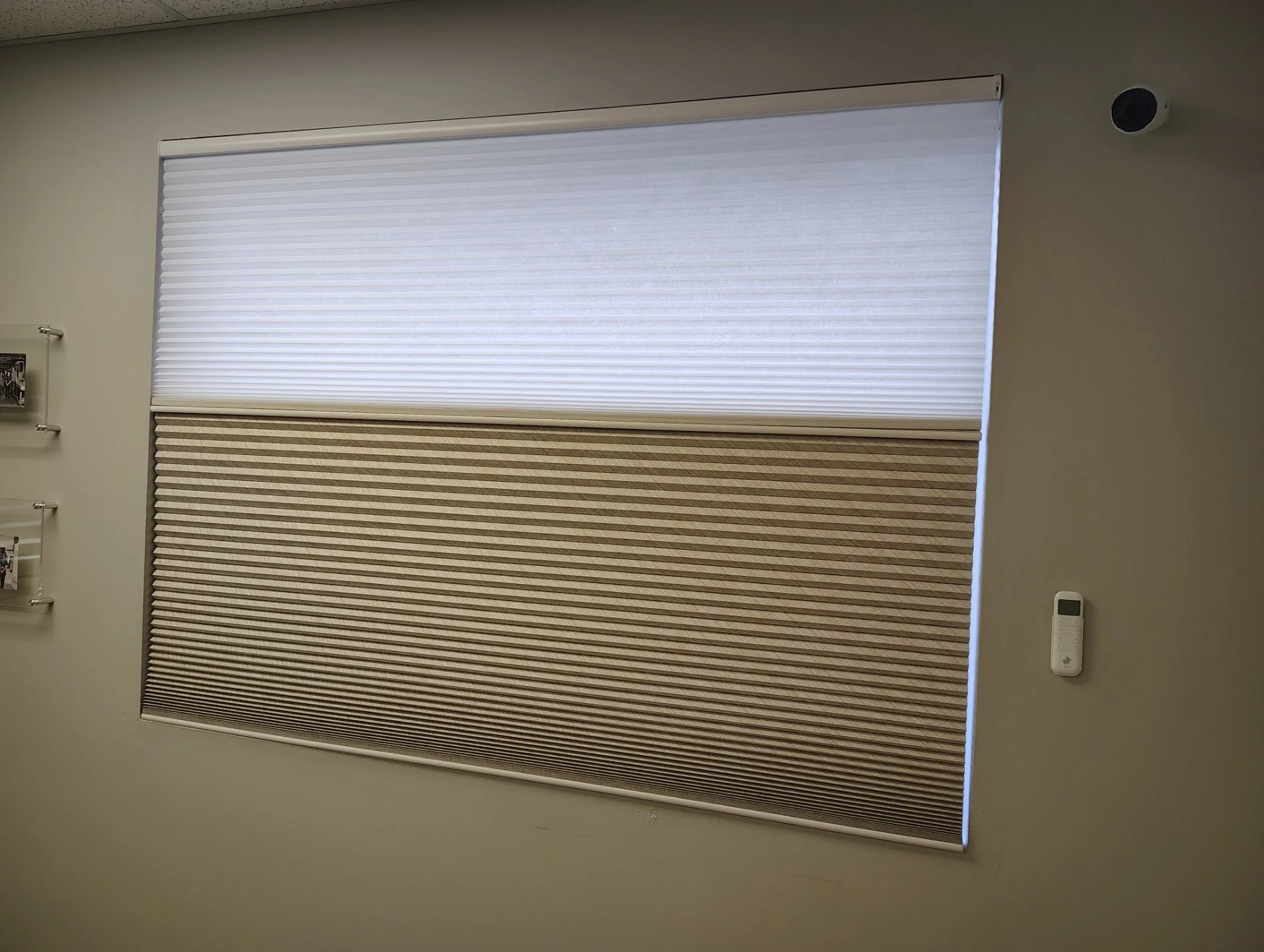 Indoor room with three large window blinds, the top is white and the bottom is beige with horizontal stripes, both covering the windows. There is a small security camera in the upper right corner, a wall-mounted thermostat in the lower right corner, 