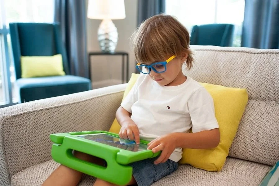 A young boy with blonde hair and blue glasses sitting on a beige couch, focused on a green electronic tablet, working on literacy skills in conjunction with AAC therapy in Washington, DC.