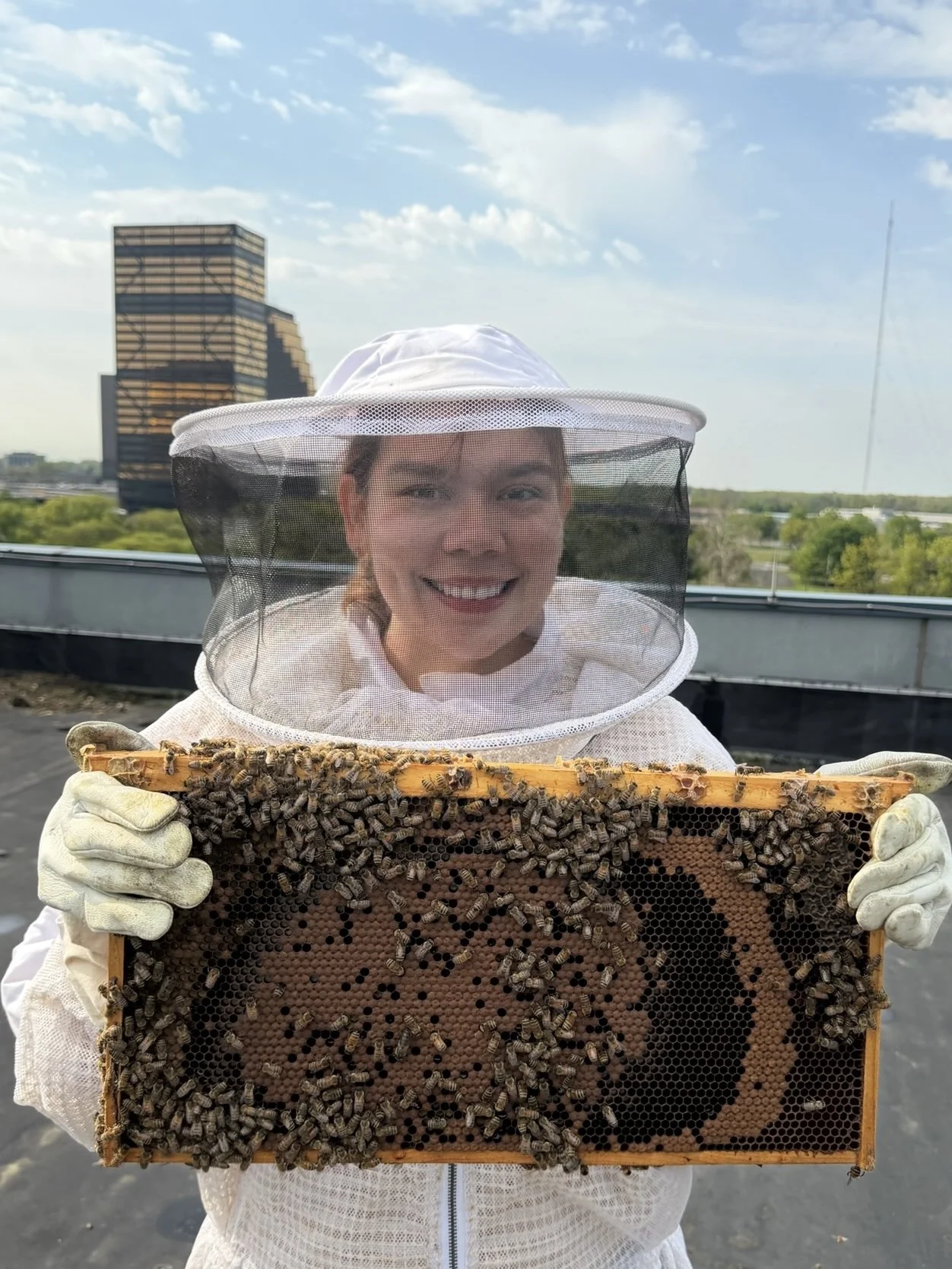 Mujer con traje de apicultora sosteniendo un panal lleno de abejas en un ambiente exterior, con edificios y árboles en el fondo.